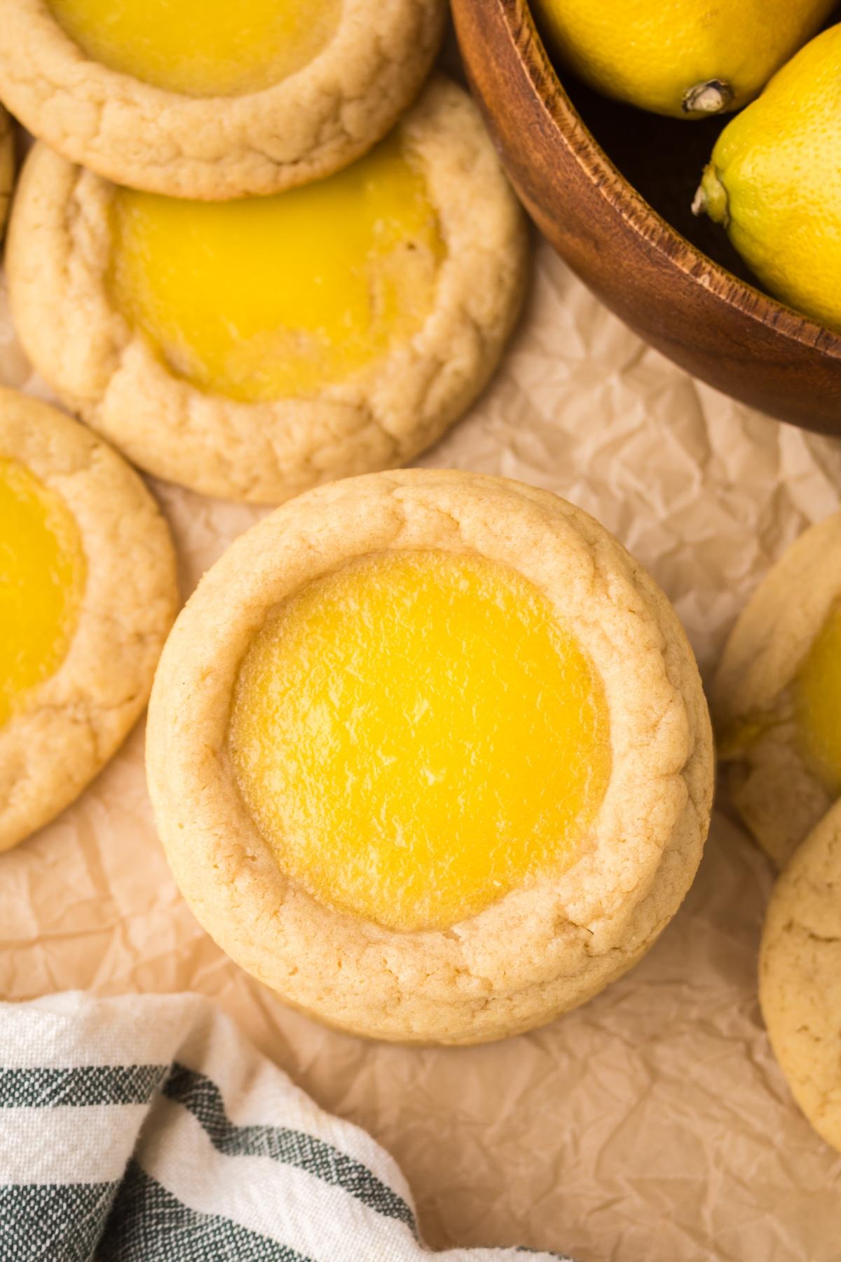 Lemon Thumbprint cookies with yellow centers on parchment paper next to whole lemons in a wooden bowl.