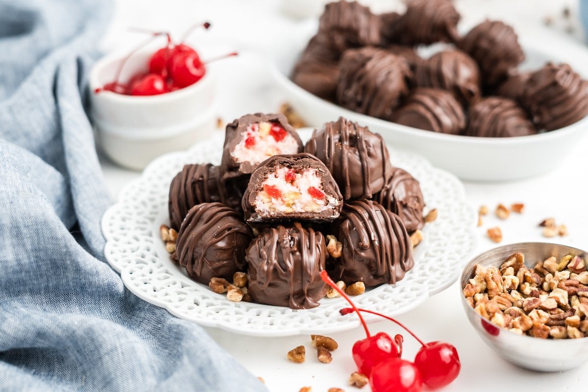 Chocolate truffles with cherry filling on a white plate, surrounded by cherries and chopped nuts—these treats evoke classic Martha Washington Candy with their rich flavors and nostalgic charm.