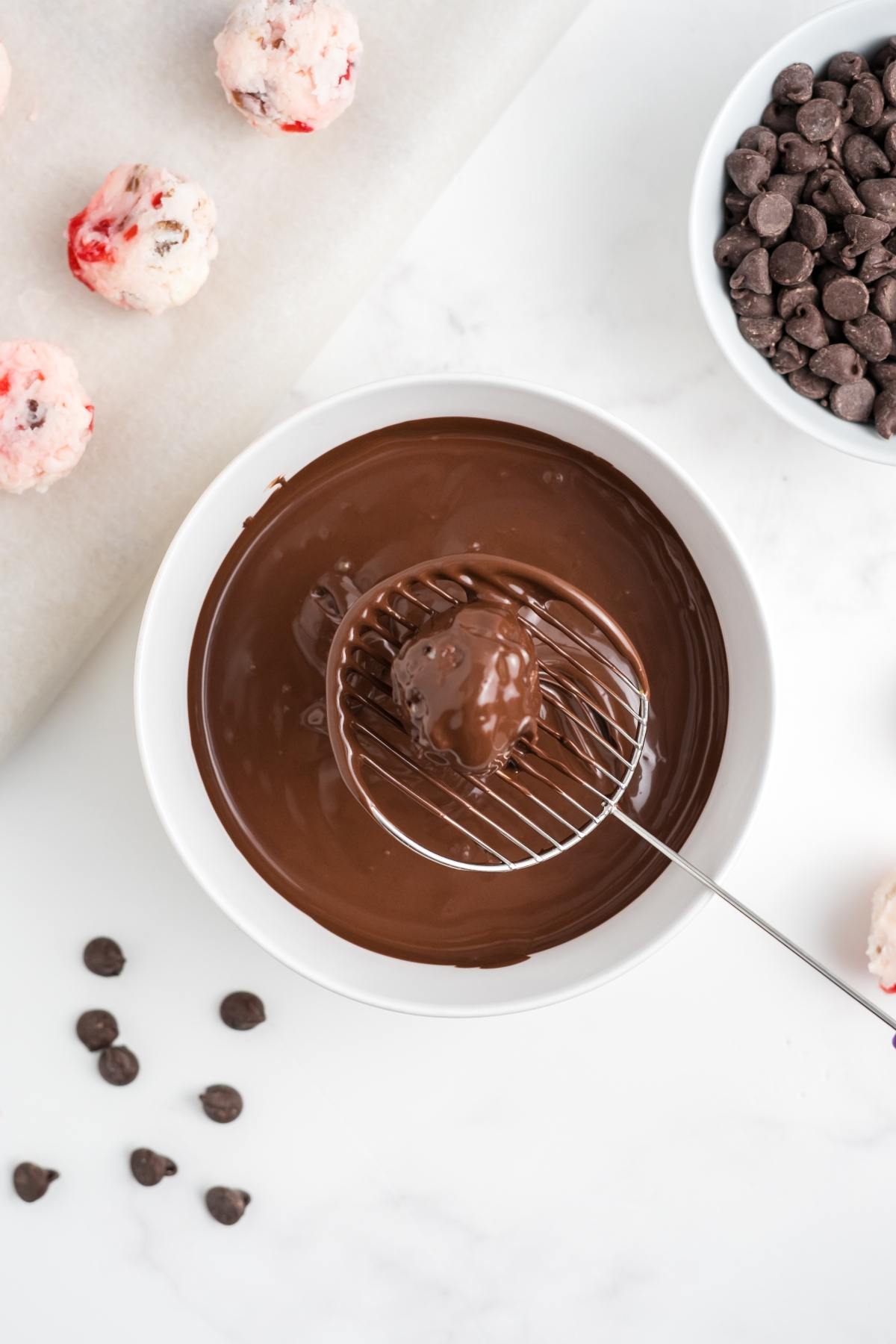 A cherry truffle being dipped in melted chocolate with chocolate chips nearby on a white surface.
