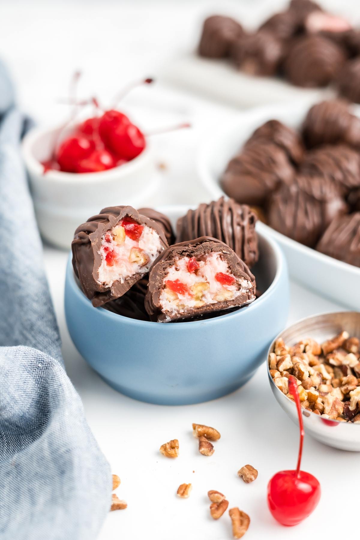 Chocolate-covered cherry martha washington candies with chopped pecans in a blue bowl, surrounded by cherries and nuts.