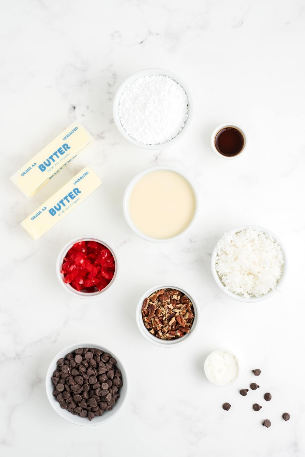 Ingredients for a dessert: butter, powdered sugar, vanilla, condensed milk, cherries, pecans, coconut, chocolate chips.