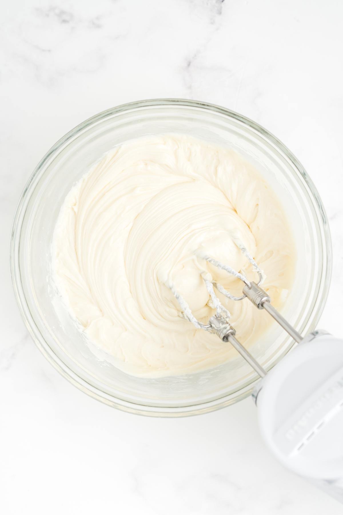 A hand mixer blending creamy white frosting in a glass bowl on a white marble surface.