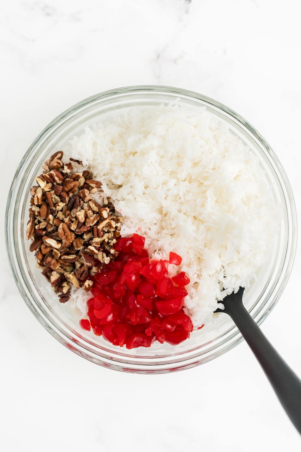 A glass bowl with shredded coconut, chopped pecans, and diced red cherries, with a black spatula inside.