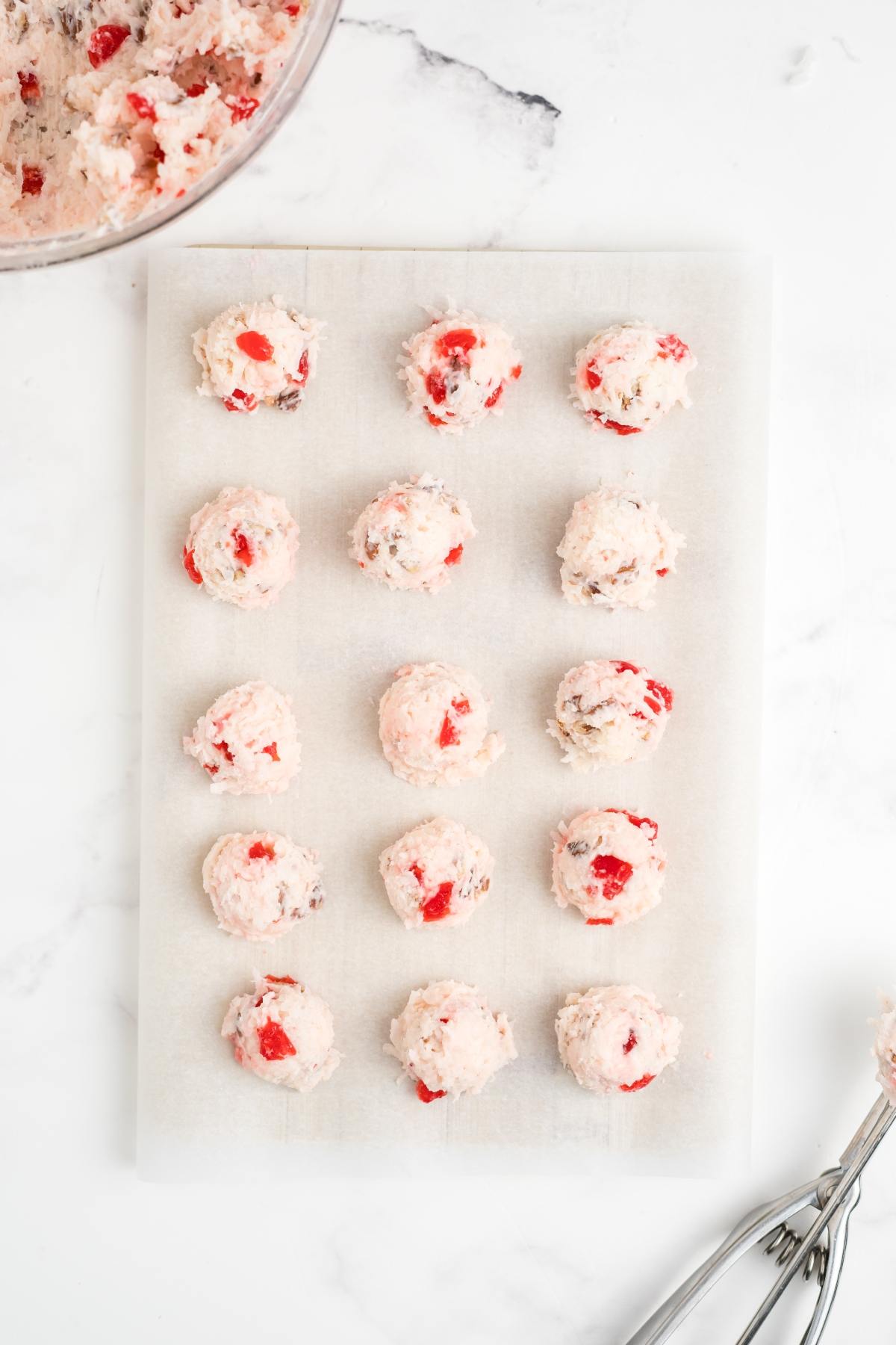 Sixteen scoops of cherry cookie dough on parchment paper, with a mixing bowl and scoop nearby on a white surface.