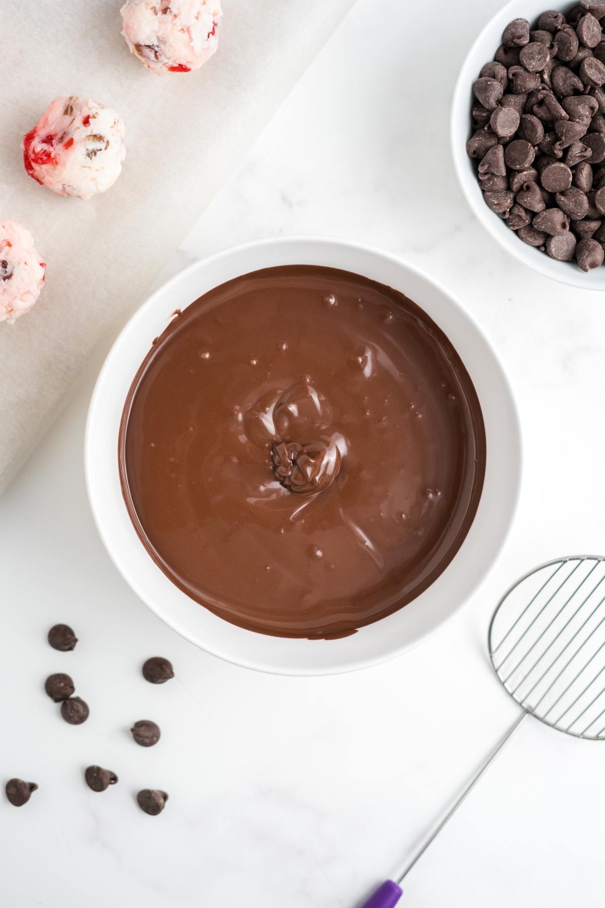 Bowl of melted chocolate with chocolate chips, cookie dough balls, and a dipping tool on a white surface.