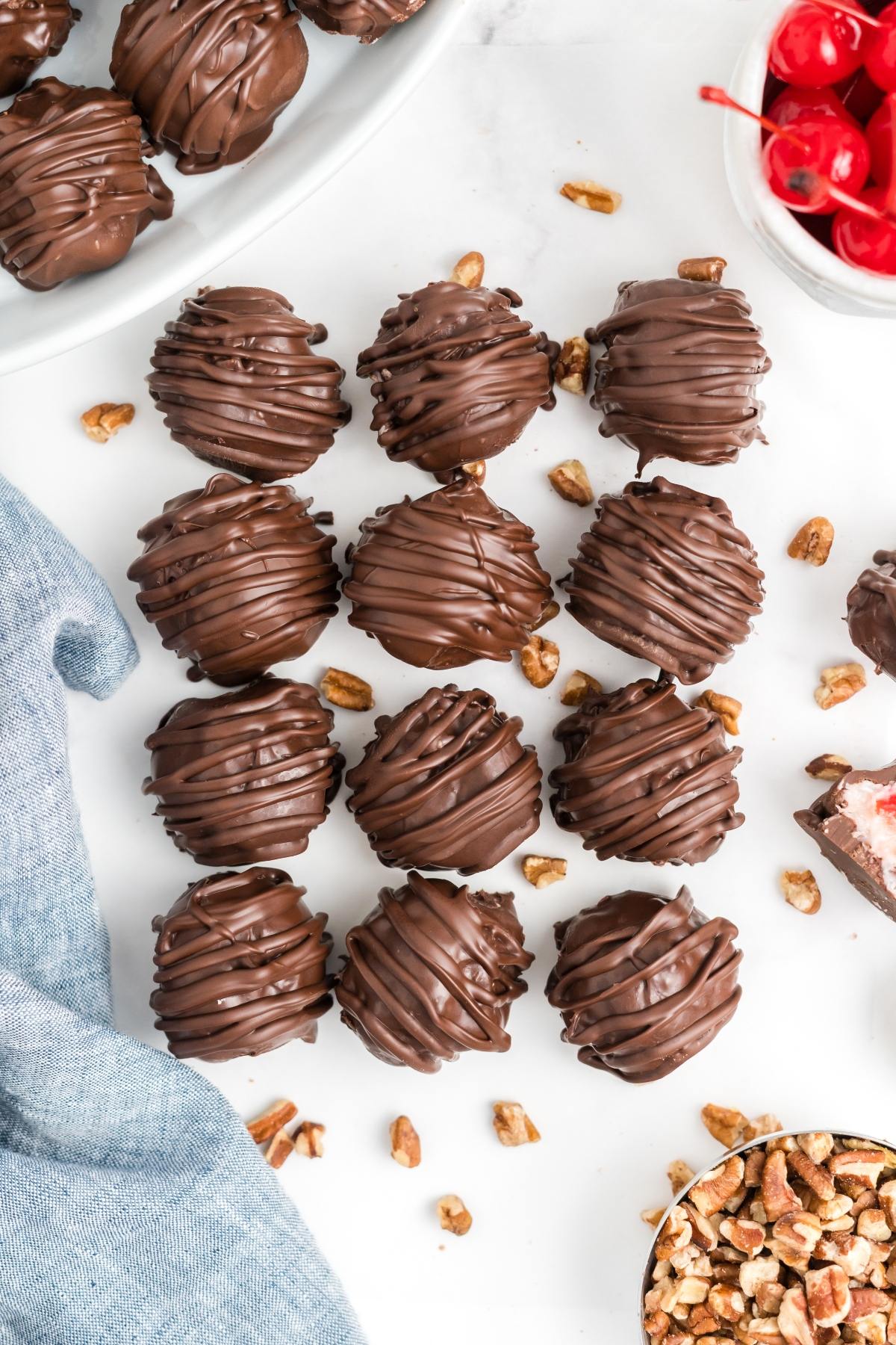 Chocolate-covered truffles arranged on a white surface, surrounded by pecans and a bowl of cherries.