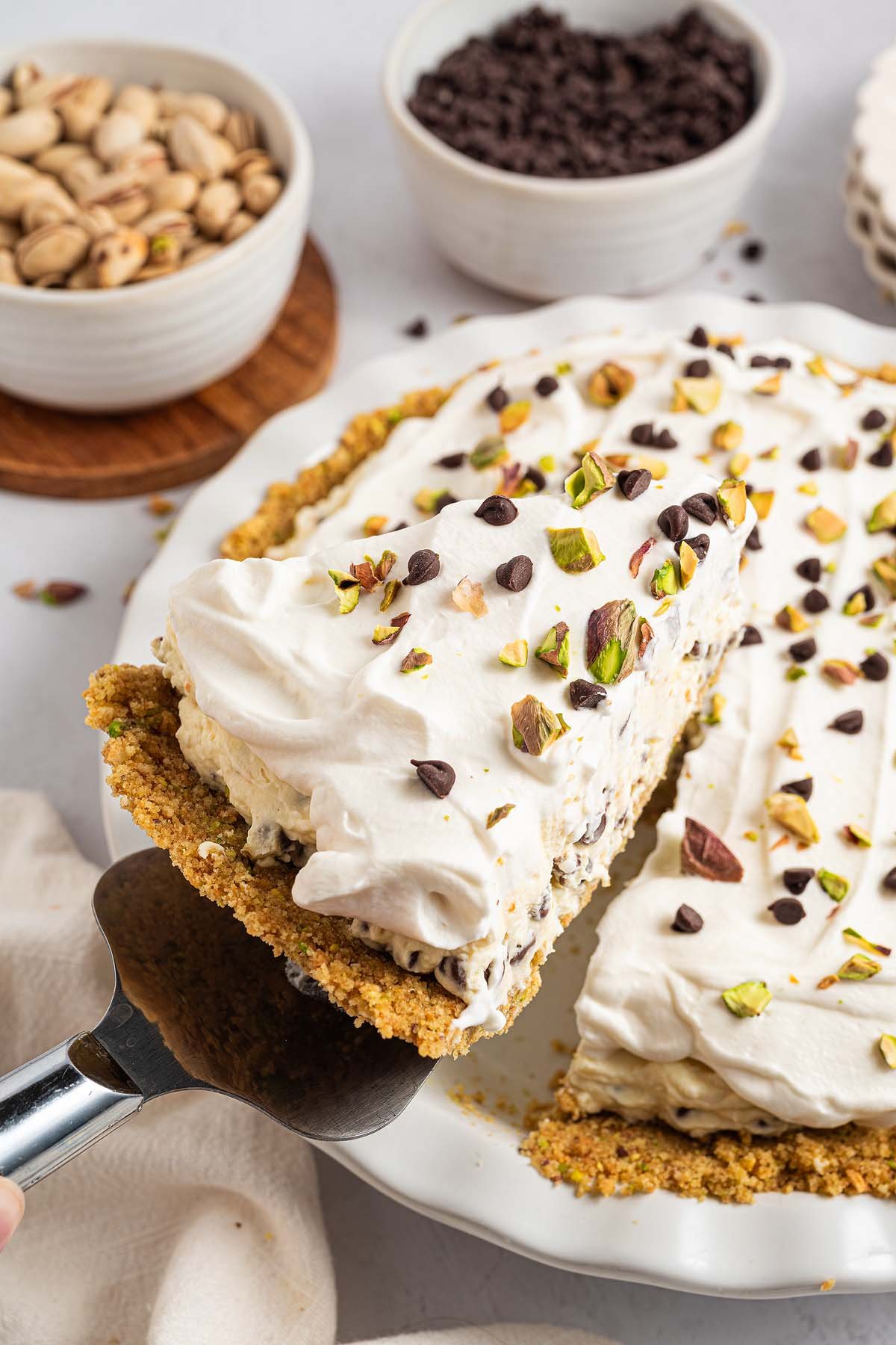 A slice of creamy Cannoli Pie with whipped topping, chocolate chips, and pistachios is being lifted from a pie dish.