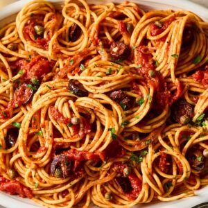 Pasta Puttanesca: Spaghetti with tomato sauce, olives, and capers served in a white oval dish, garnished with fresh herbs.