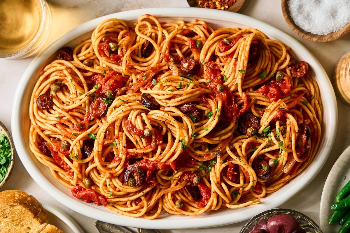 A platter of Pasta Puttanesca&mdash;spaghetti tossed with tomato sauce, olives, and herbs&mdash;is surrounded by various small dishes.