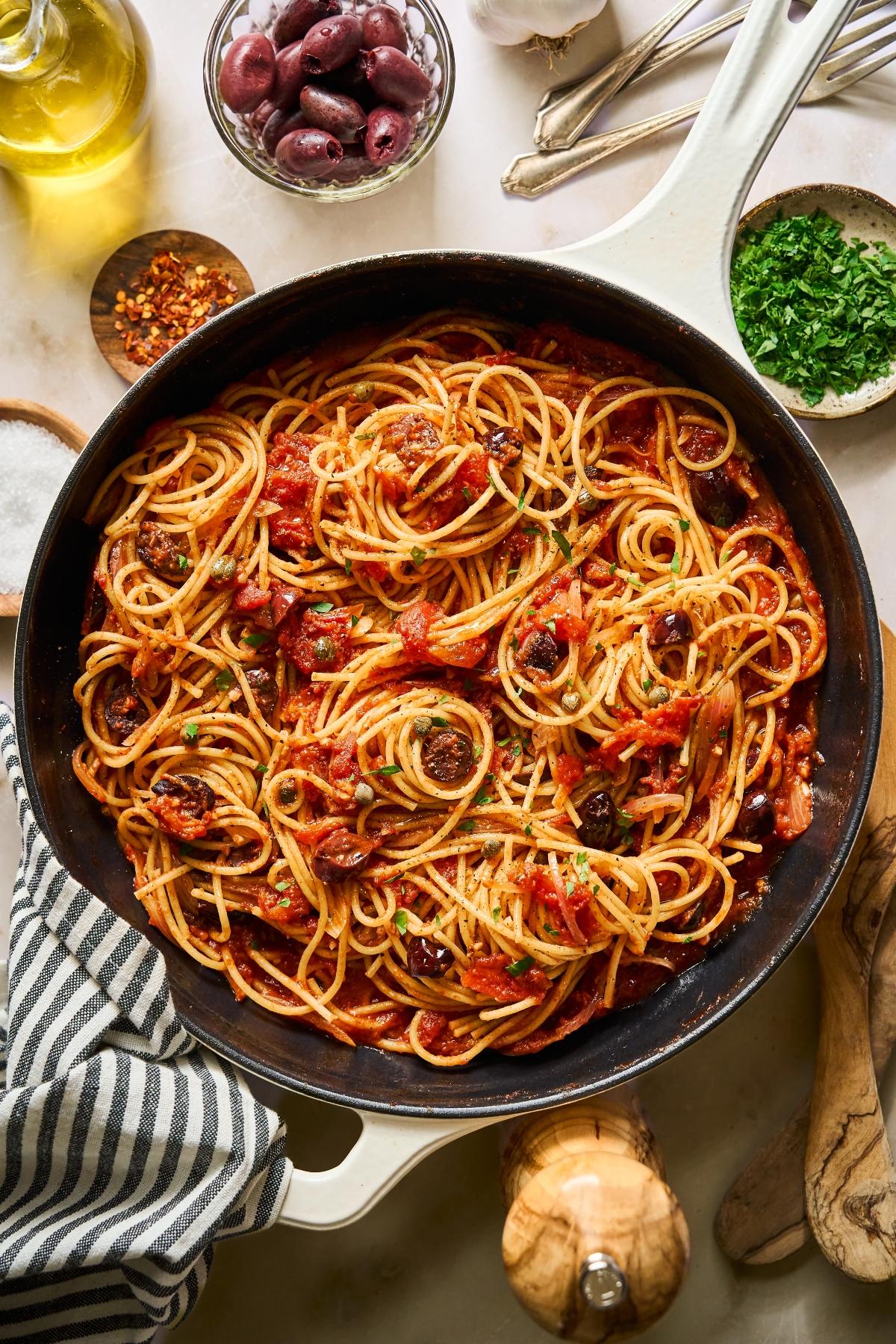 Spaghetti with tomato sauce, olives, and herbs in a pan, surrounded by fresh ingredients on a counter.