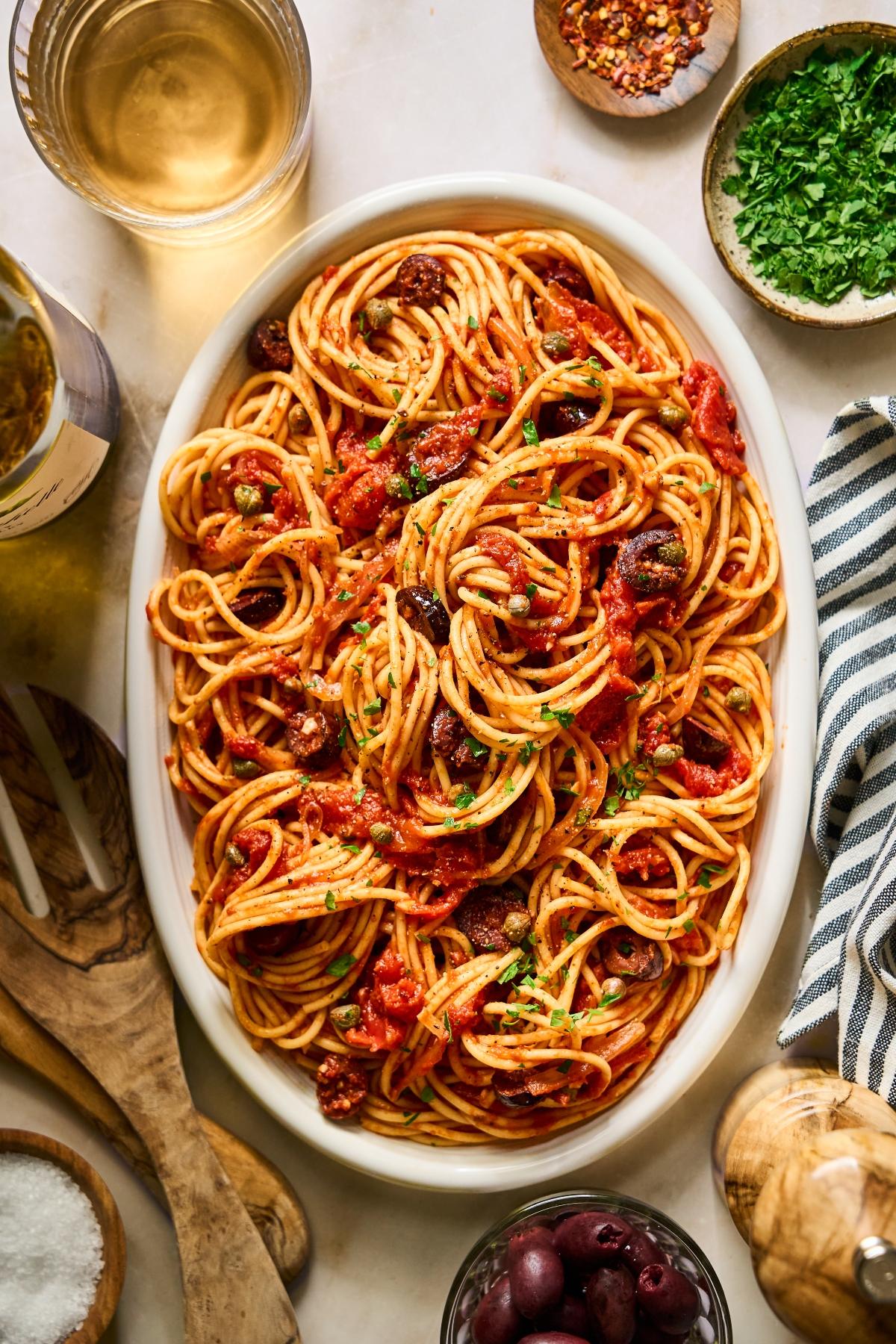 Oval platter of spaghetti with tomato sauce, olives, and herbs, surrounded by wine, spices, and fresh parsley.
