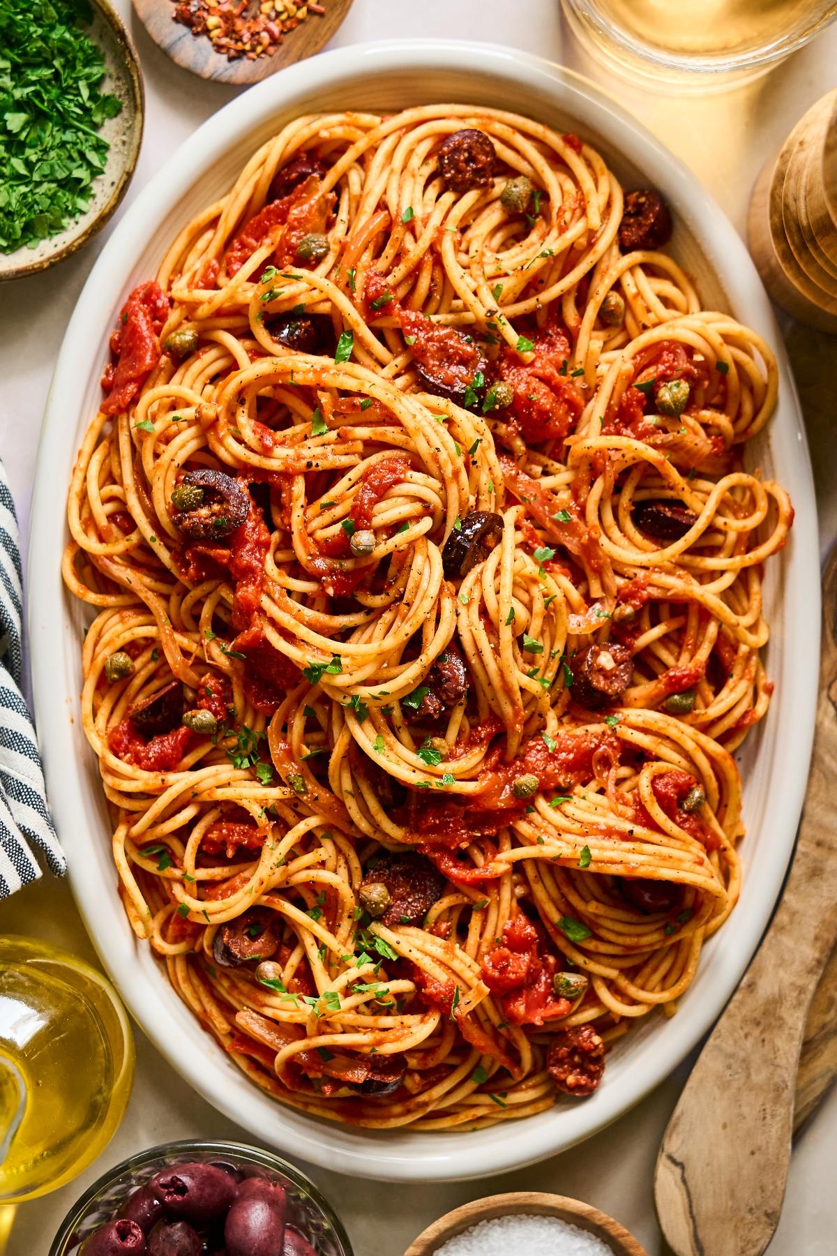 Oval dish of spaghetti with tomato sauce, olives, capers, and herbs, surrounded by small bowls of ingredients.