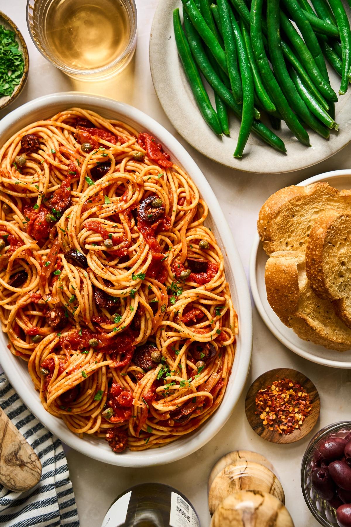 Oval dish of spaghetti with tomato sauce and olives, green beans, toasted bread, and a glass of white wine.