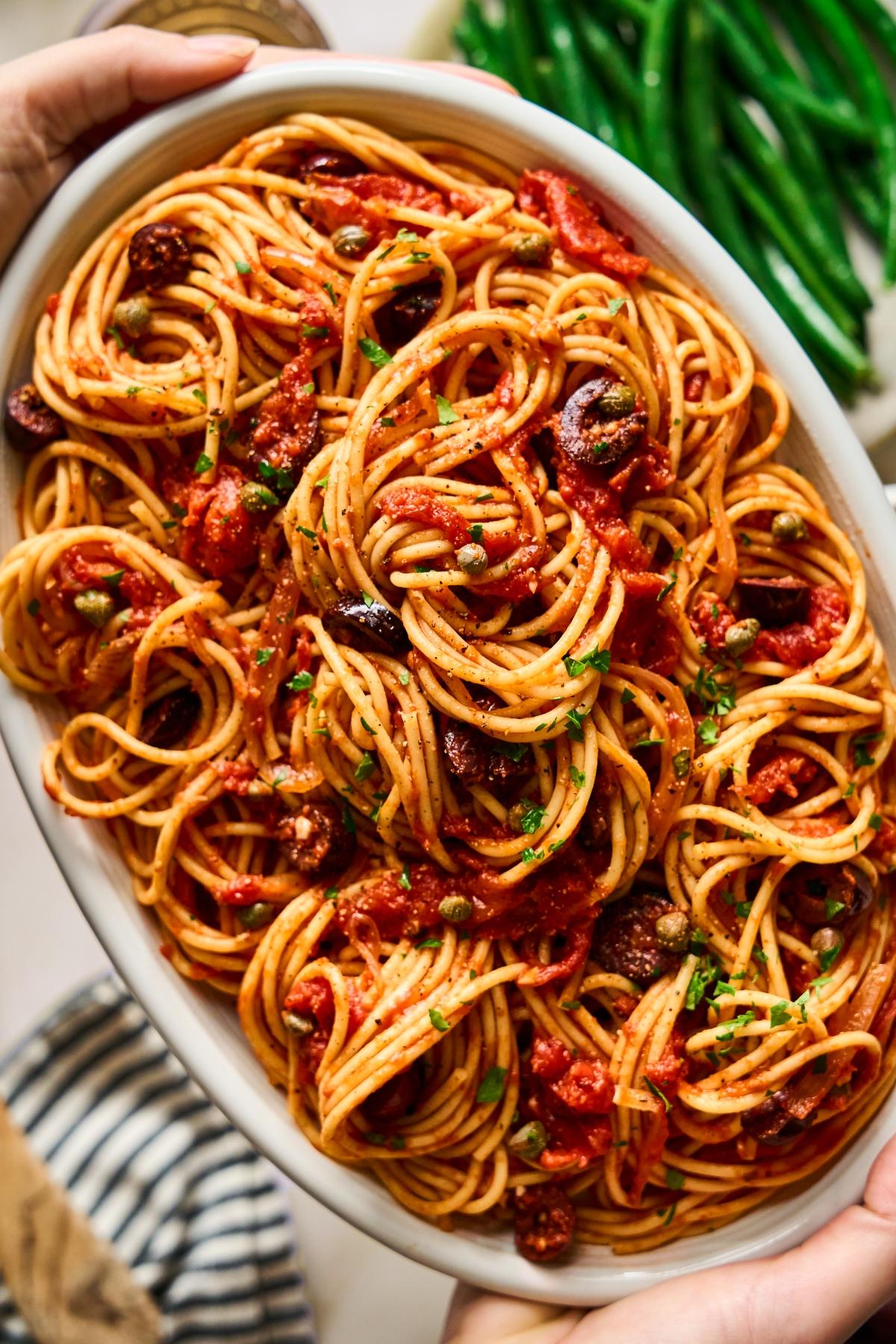 A dish of spaghetti with tomato sauce, olives, and capers, garnished with herbs, served on a white platter.