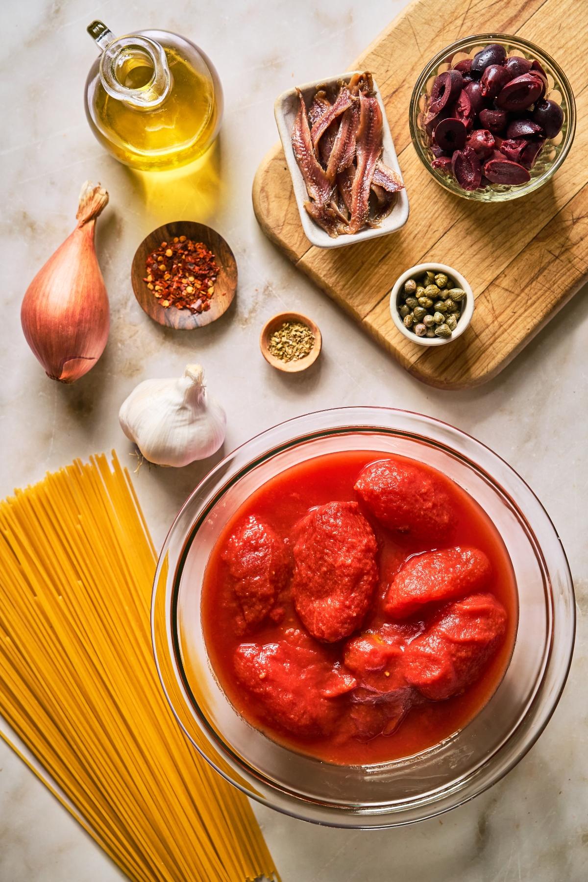 Top view of pasta, canned tomatoes, garlic, shallot, olives, anchovies, capers, spices, and olive oil on a counter.