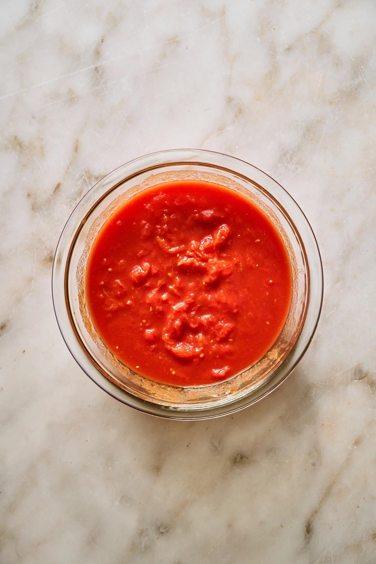 A glass bowl of chunky tomato sauce on a marble surface.
