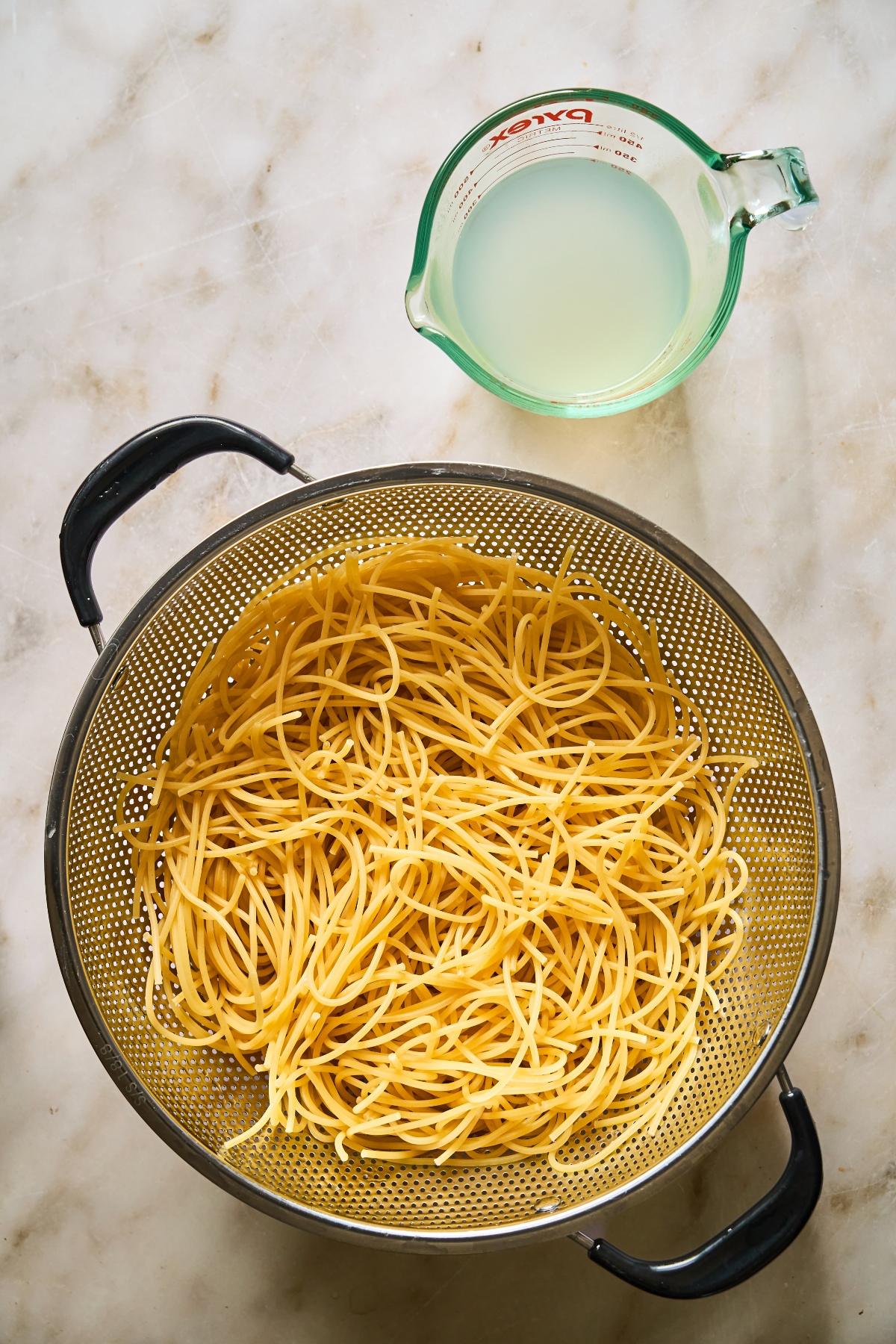 Cooked spaghetti in a colander next to a glass measuring cup filled with pasta water on a marble surface.