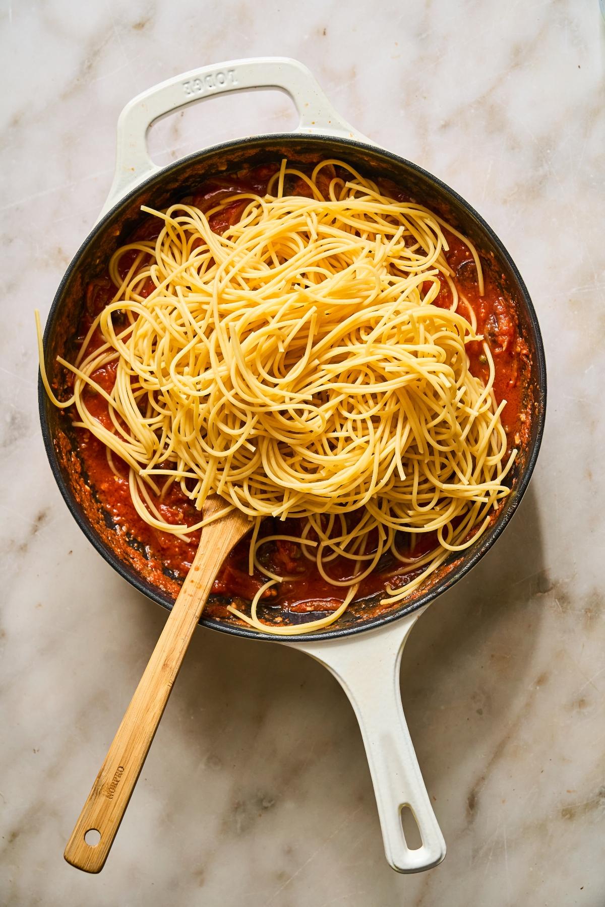 A white pan with cooked spaghetti and tomato sauce, stirred with a wooden spoon on a marble surface.