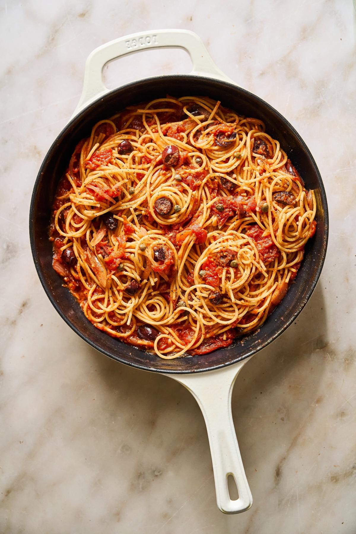 Spaghetti with tomato sauce and olives in a white skillet on a marble surface.