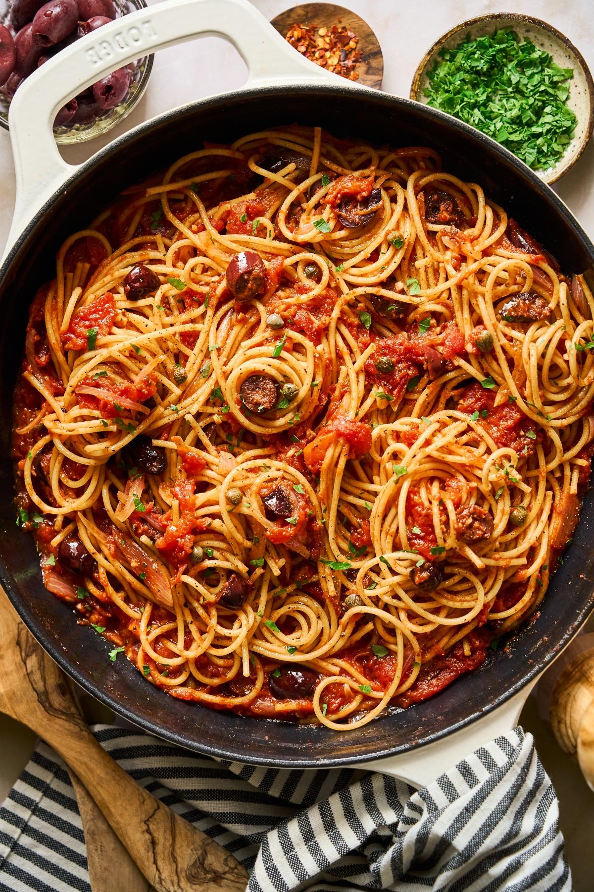 Spaghetti with tomato sauce, olives, and herbs in a large black pan, surrounded by bowls of parsley and chili flakes.