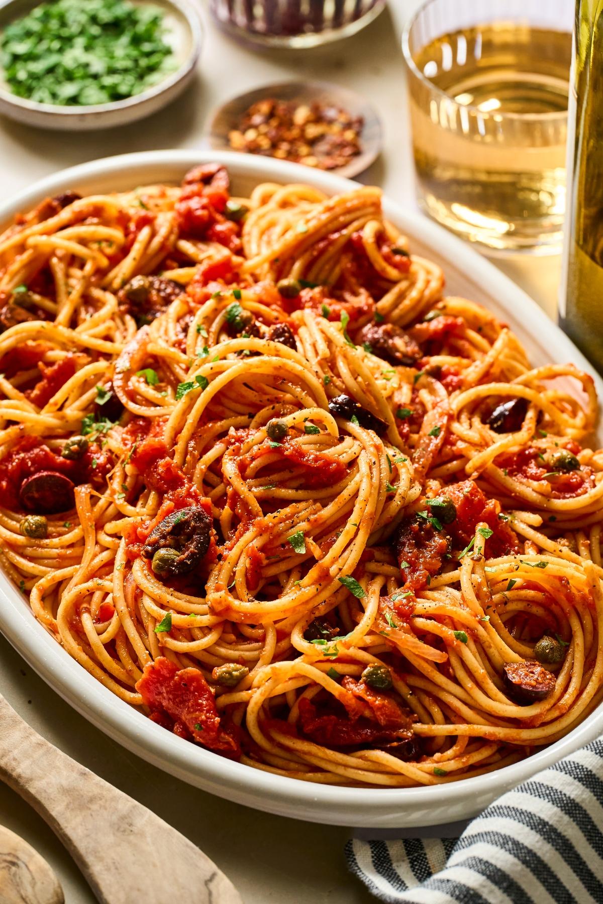 A plate of spaghetti with tomato sauce, olives, and herbs, with wine and seasonings in the background.