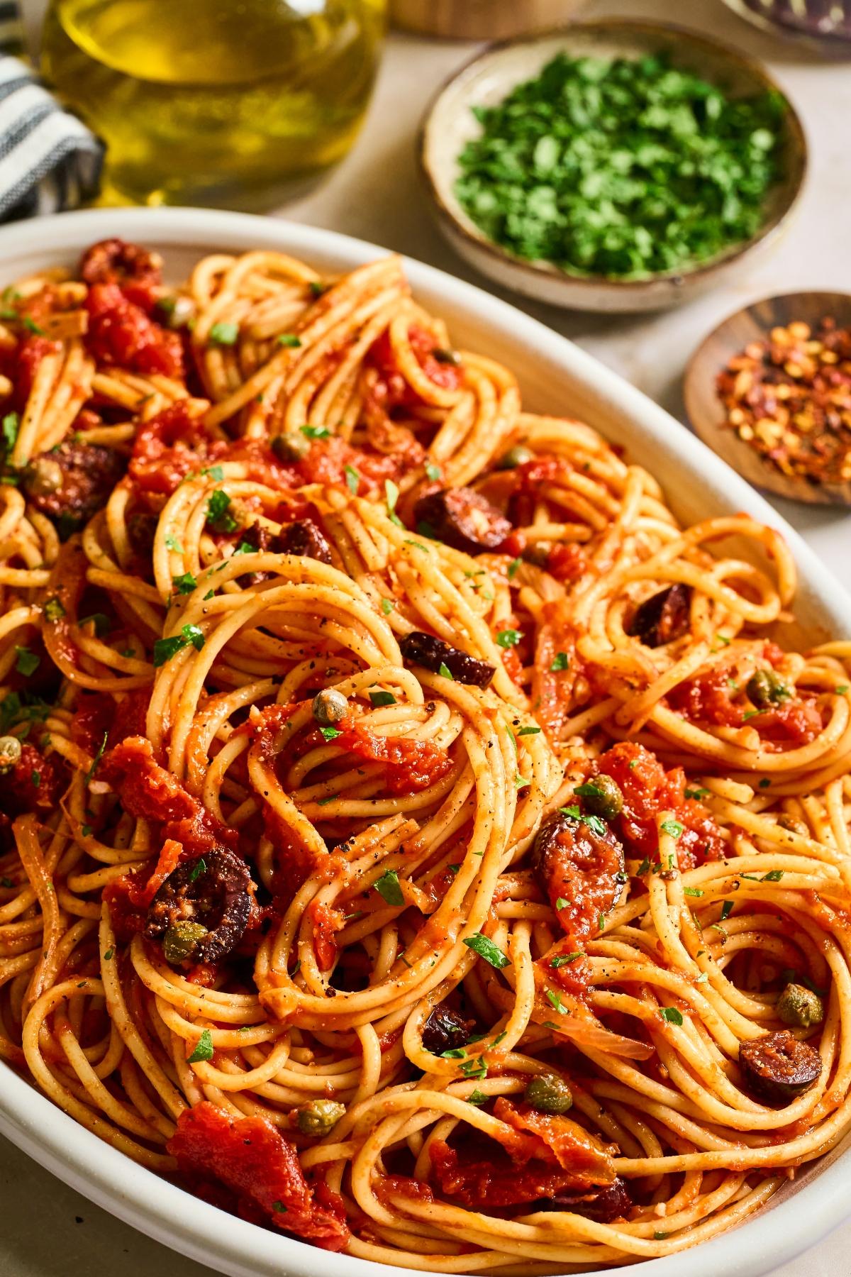 A platter of spaghetti with tomato sauce, olives, capers, and parsley, with seasonings in small bowls nearby.