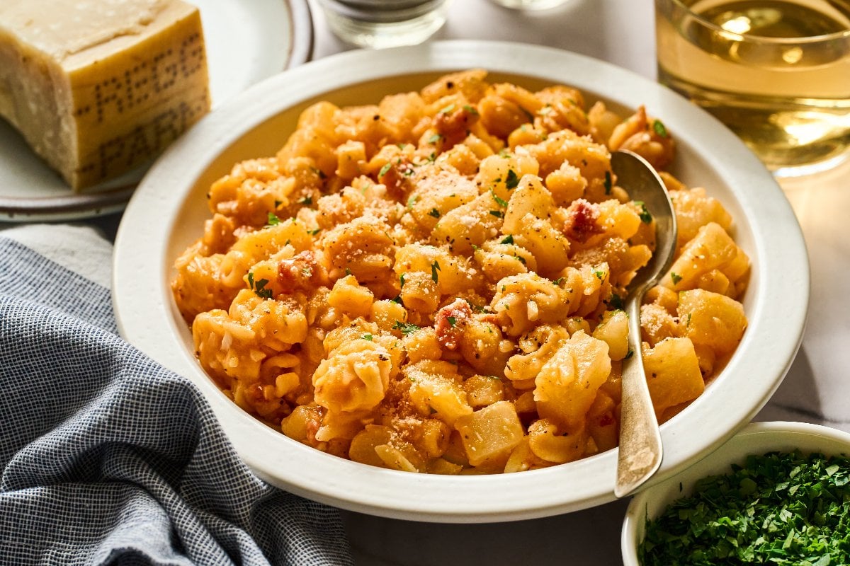 A bowl of Pasta e Patate with tomato sauce and cheese, garnished with herbs, sits beside a spoon and a block of cheese.