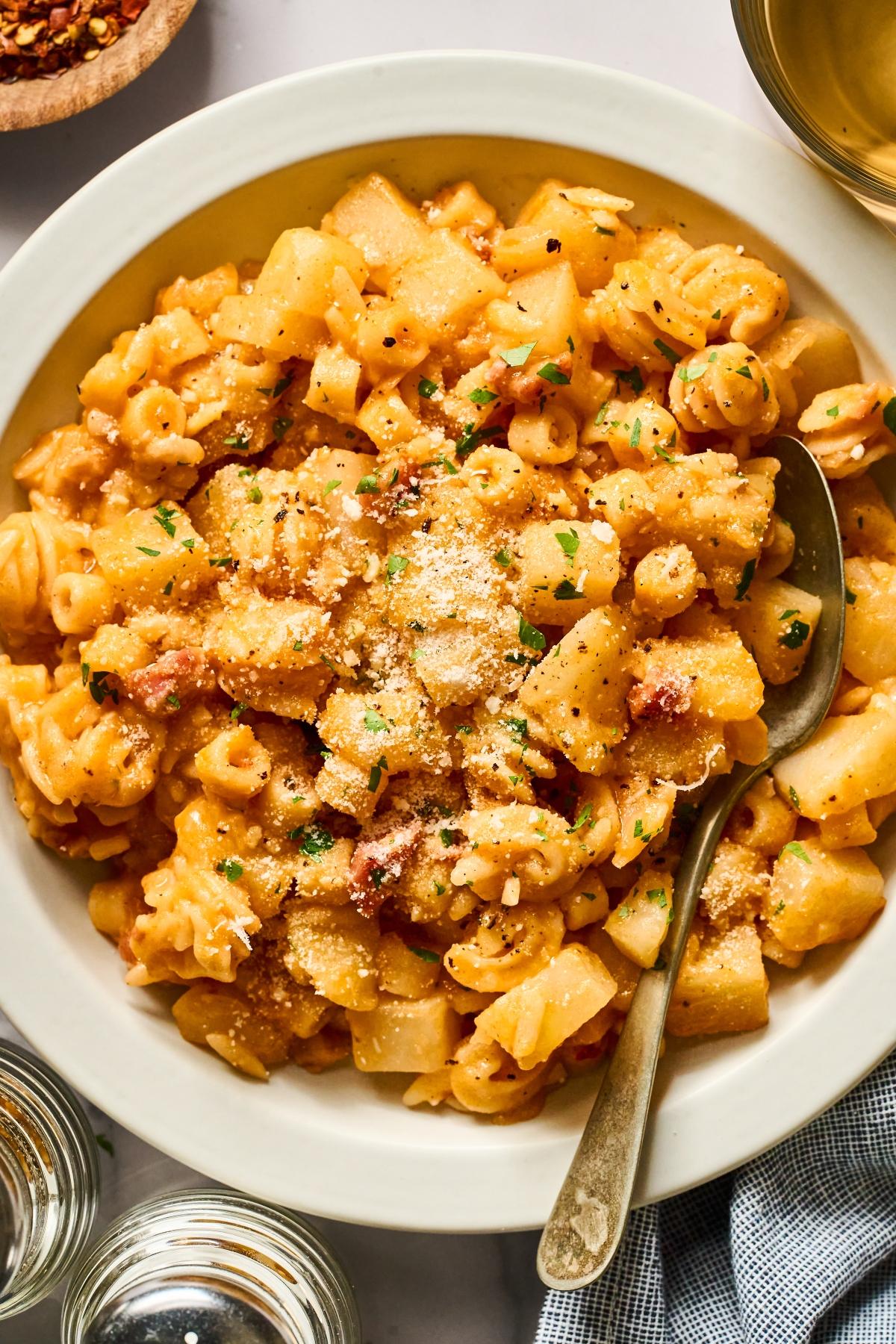 A bowl of pasta with potatoes, sprinkled with grated cheese and herbs, with a spoon on the side.