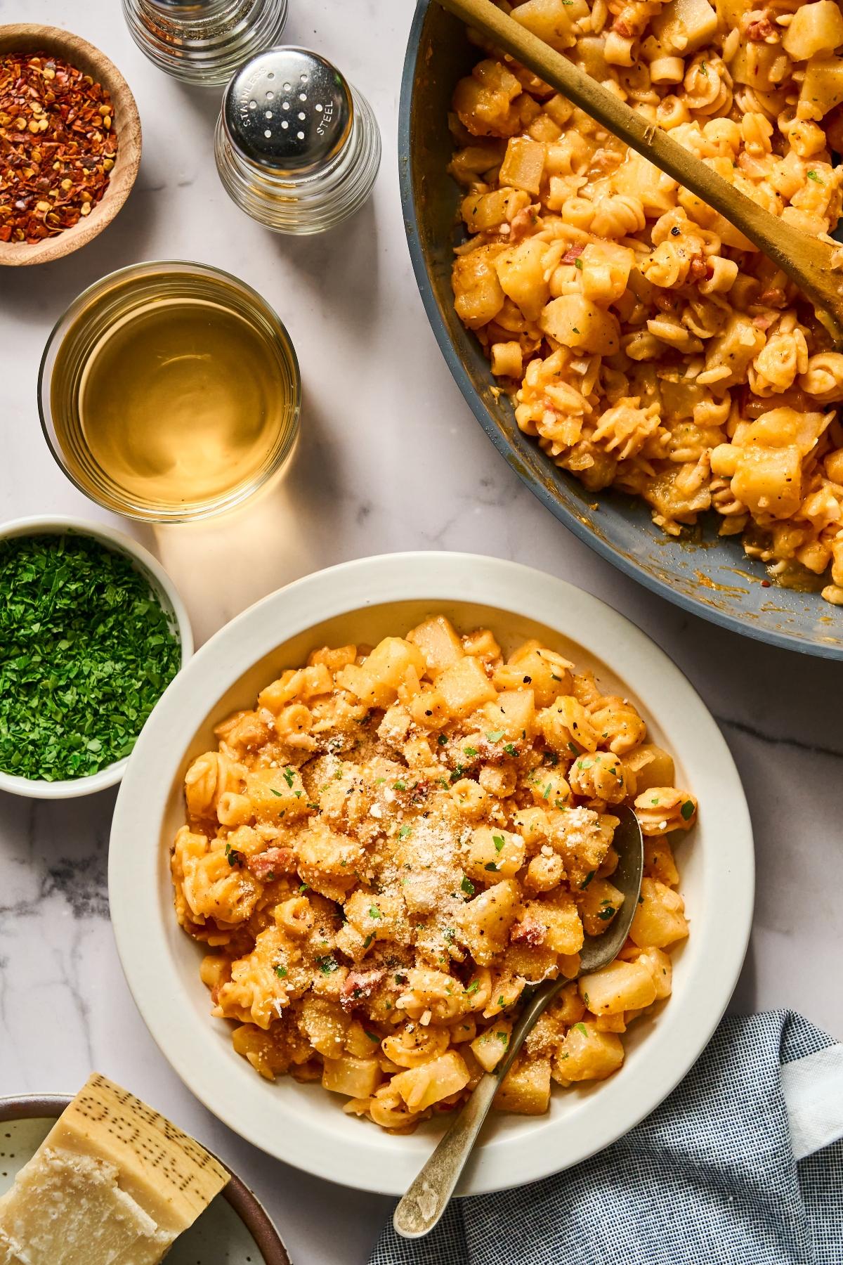 A bowl of creamy pasta with potatoes, cheese, and herbs next to a skillet, wine, and seasonings on a table.