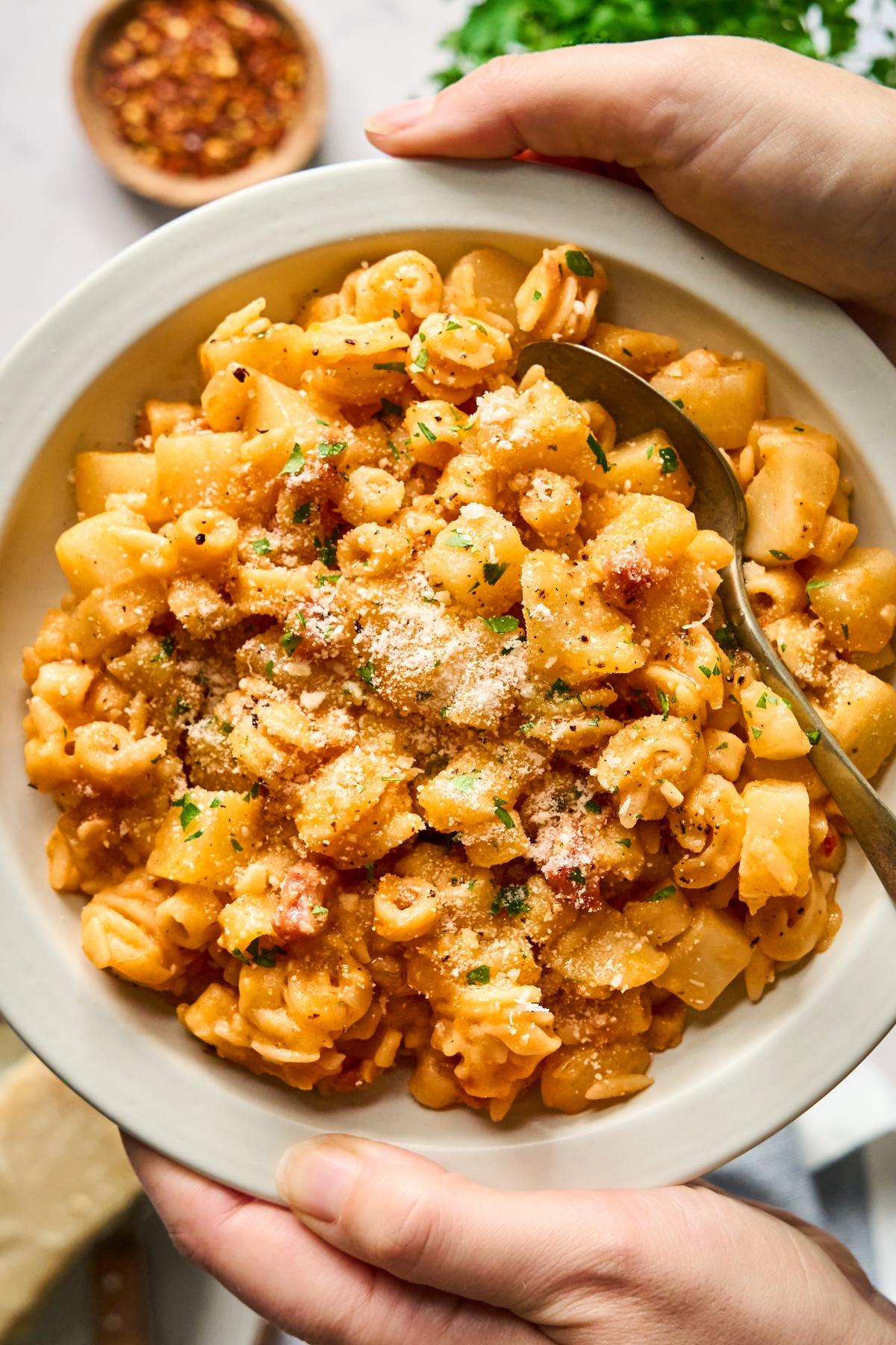 A bowl of pasta and potatoes in a tomato sauce, topped with grated cheese and parsley, held by two hands.
