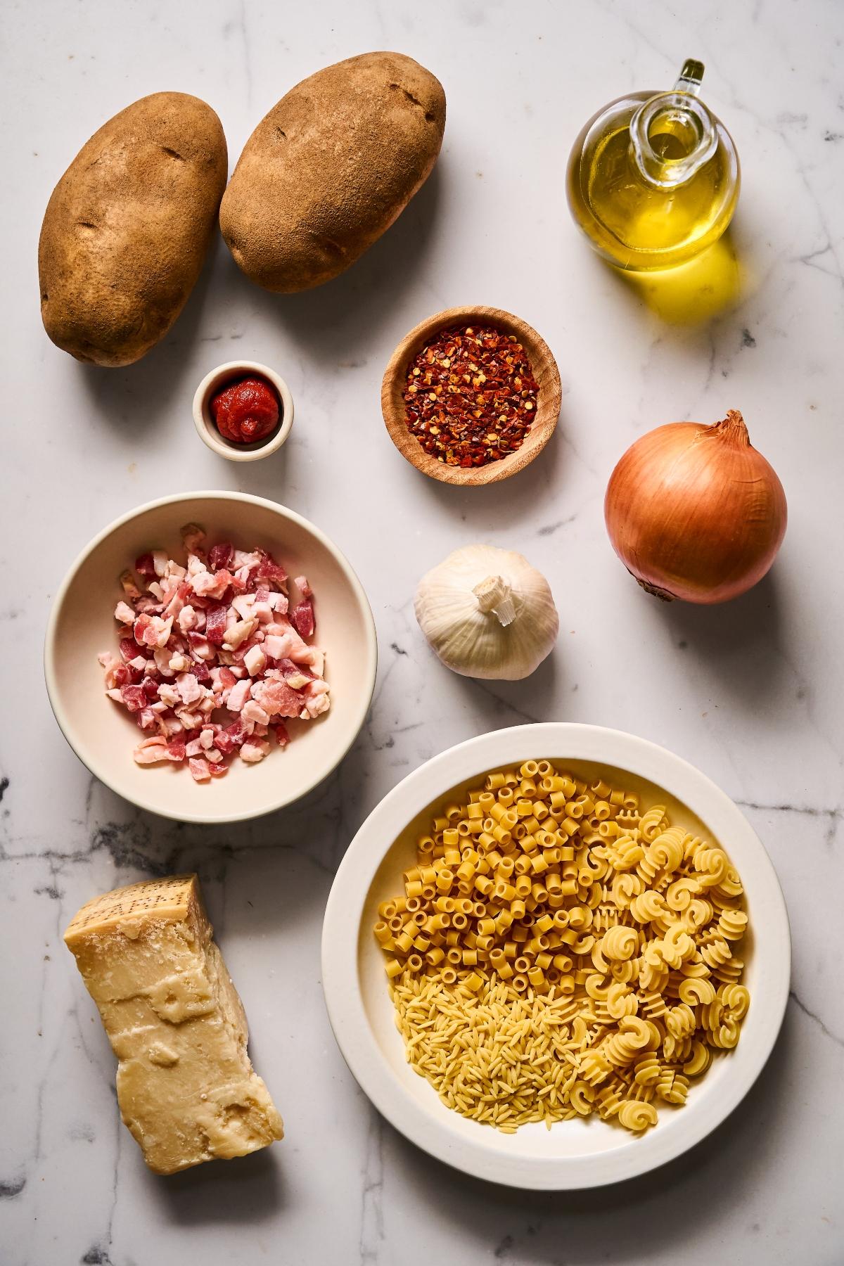 Assorted pasta, potatoes, pancetta, parmesan, onion, garlic, olive oil, tomato paste, and chili flakes on a counter.