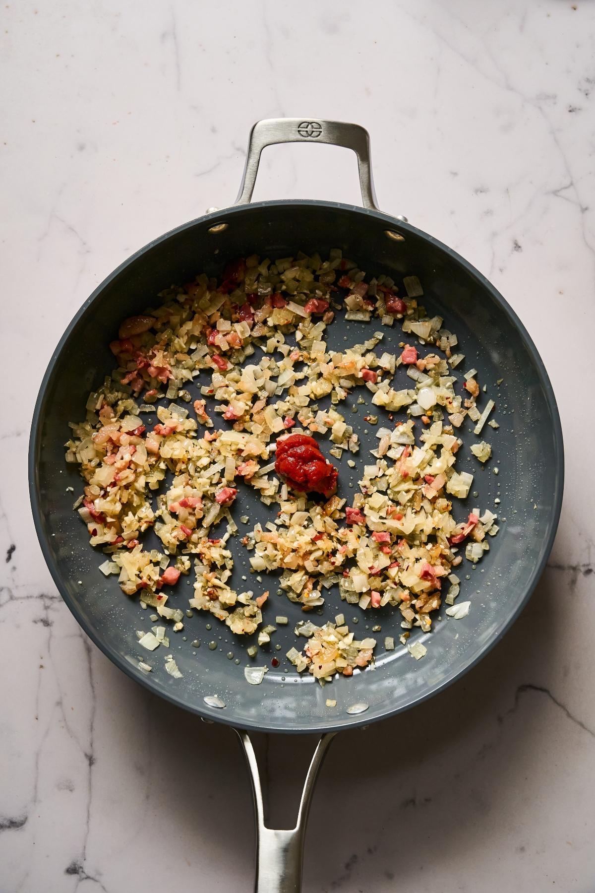 Chopped onions, garlic, and bacon sautéing in a pan with a dollop of tomato paste in the center.