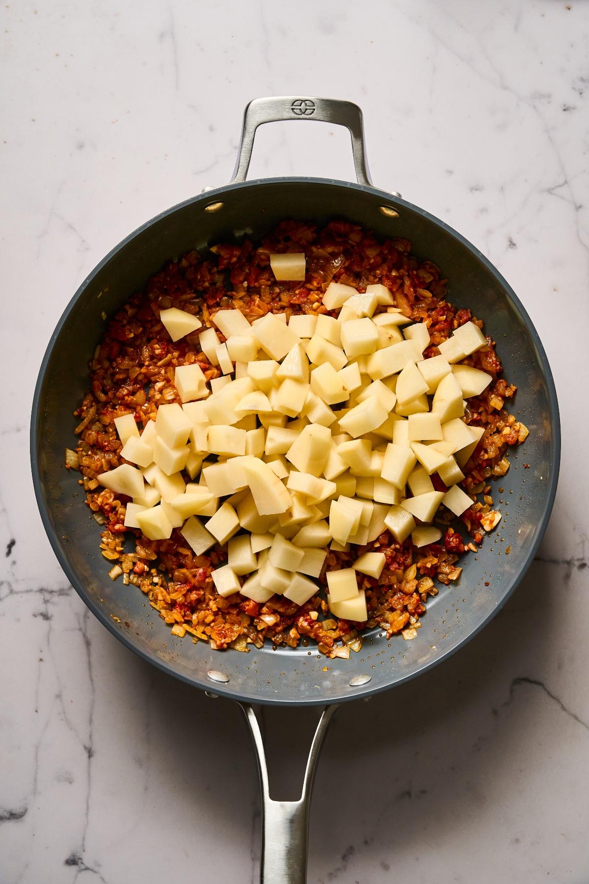 A skillet with diced potatoes on top of sautéed onions, garlic, and spices on a marble counter.