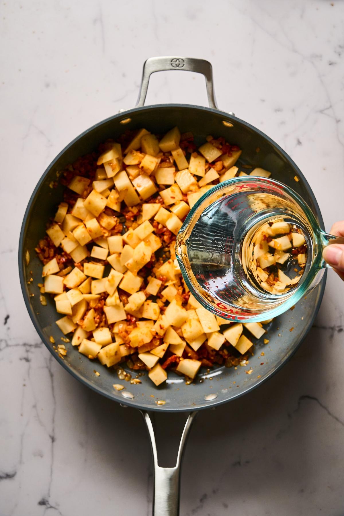 A hand pours water into a skillet with diced potatoes and seasonings on a marble countertop.