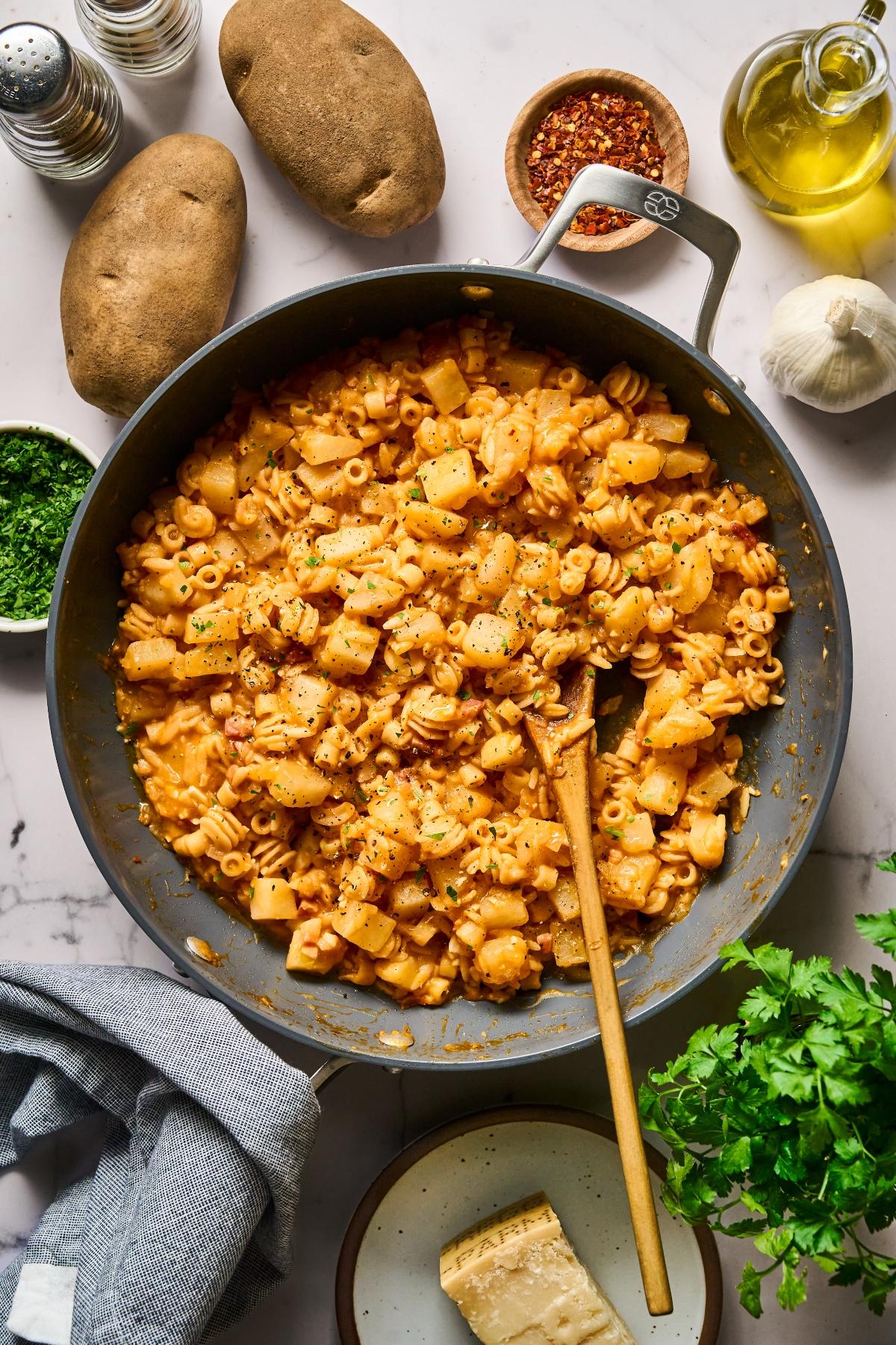 A skillet of pasta with potatoes in a creamy tomato sauce, surrounded by potatoes, garlic, cheese, and herbs.