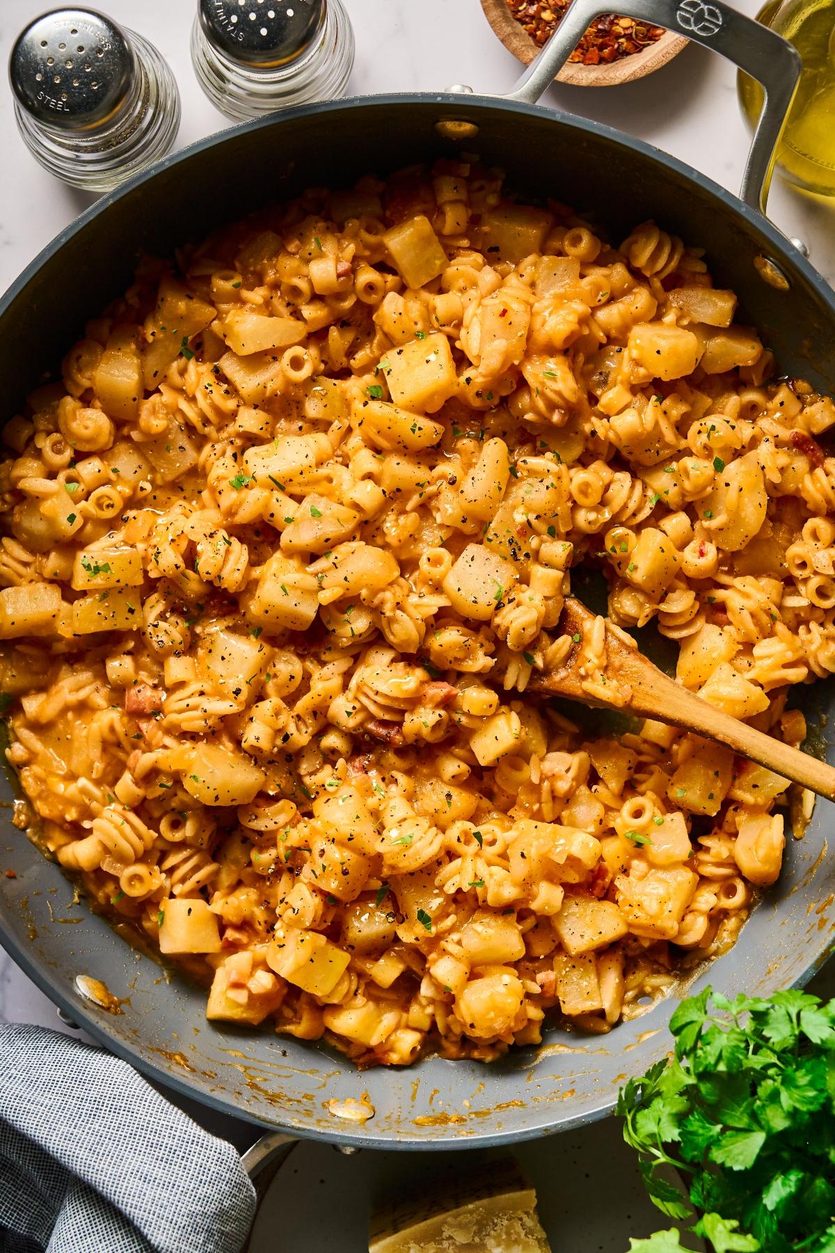 Pasta with potatoes in a creamy tomato sauce in a skillet, topped with herbs and black pepper, with a wooden spoon.