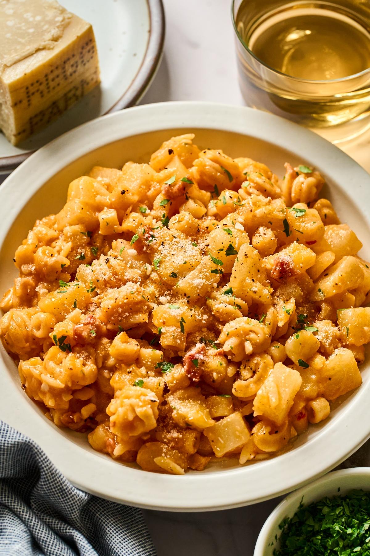A bowl of pasta with potatoes and sauce, topped with grated cheese, next to a glass of white wine.