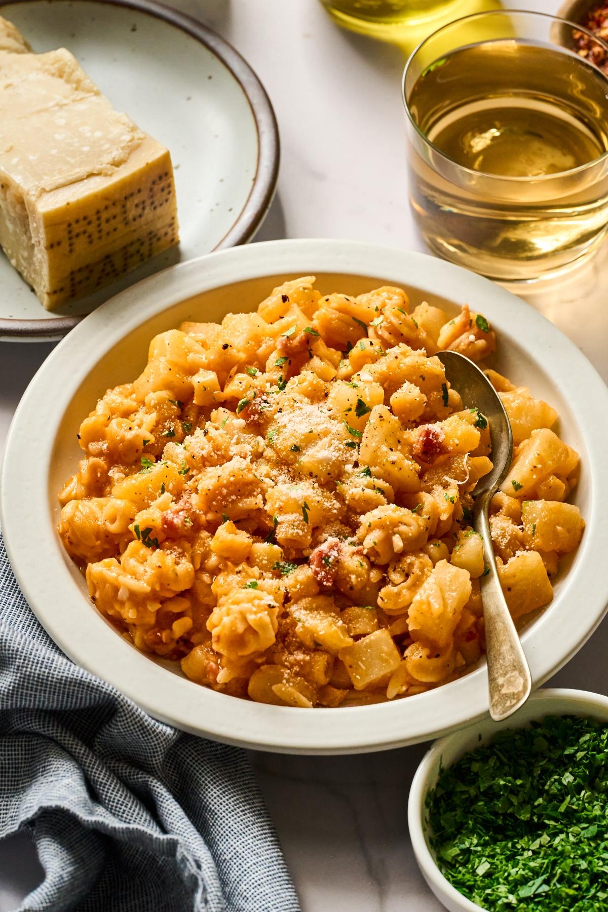 A bowl of pasta with tomato sauce, sprinkled with cheese and herbs, next to cheese, wine, and parsley.