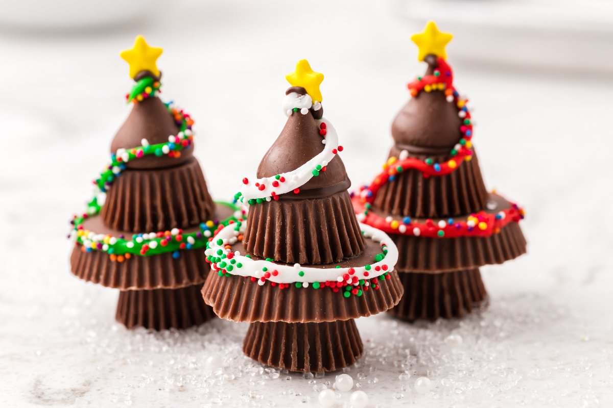 Three Peanut Butter Cup Christmas Trees are made by stacking chocolate treats, then decorating with icing and colorful sprinkles for a festive holiday dessert.