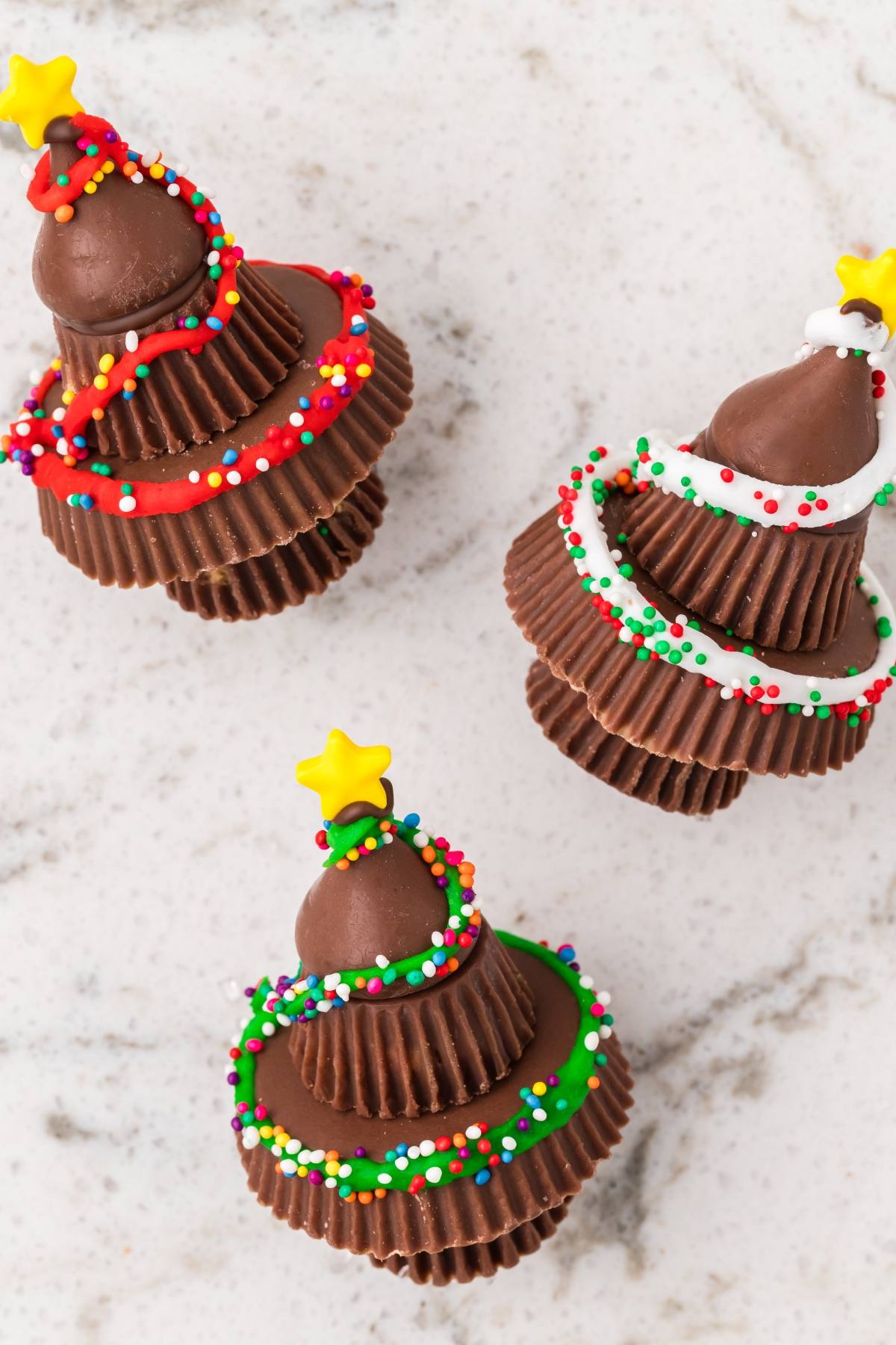 Three chocolate Christmas tree cupcakes decorated with colorful sprinkles and yellow star toppers on a white surface.