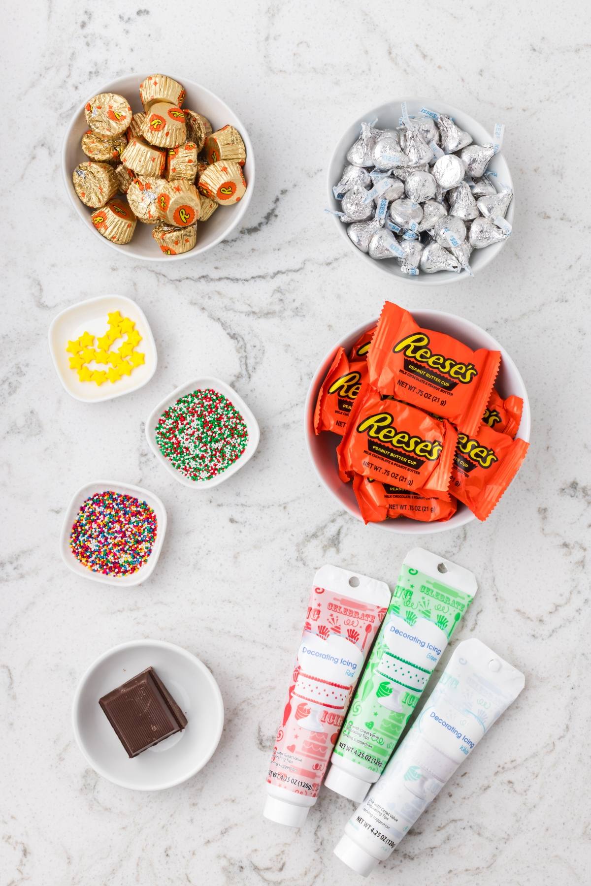 Candy, sprinkles, chocolate, and decorating gel tubes arranged in bowls on a white marble surface.