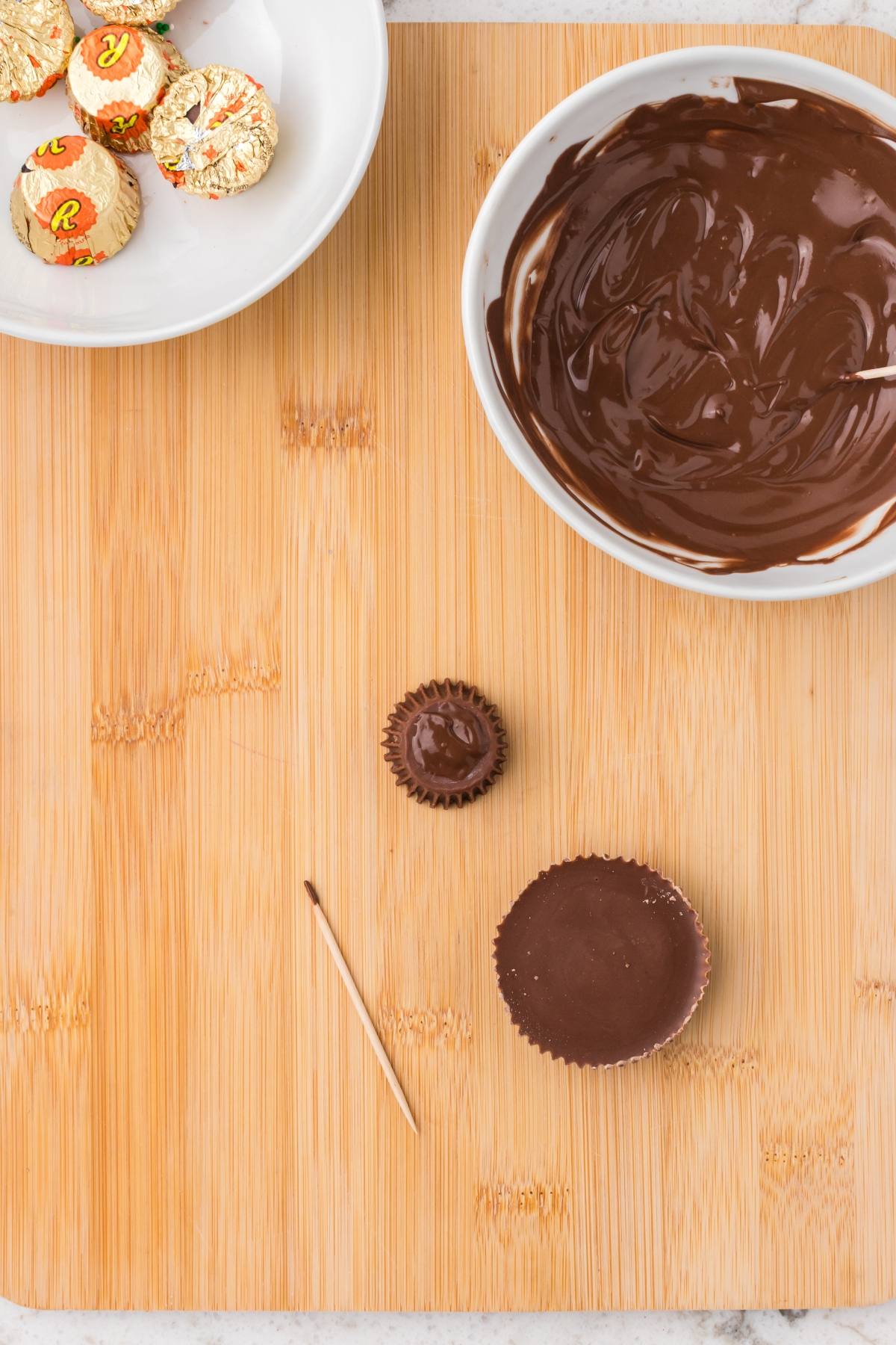 Unwrapped peanut butter cups, a bowl of melted chocolate, and a toothpick on a wooden surface.