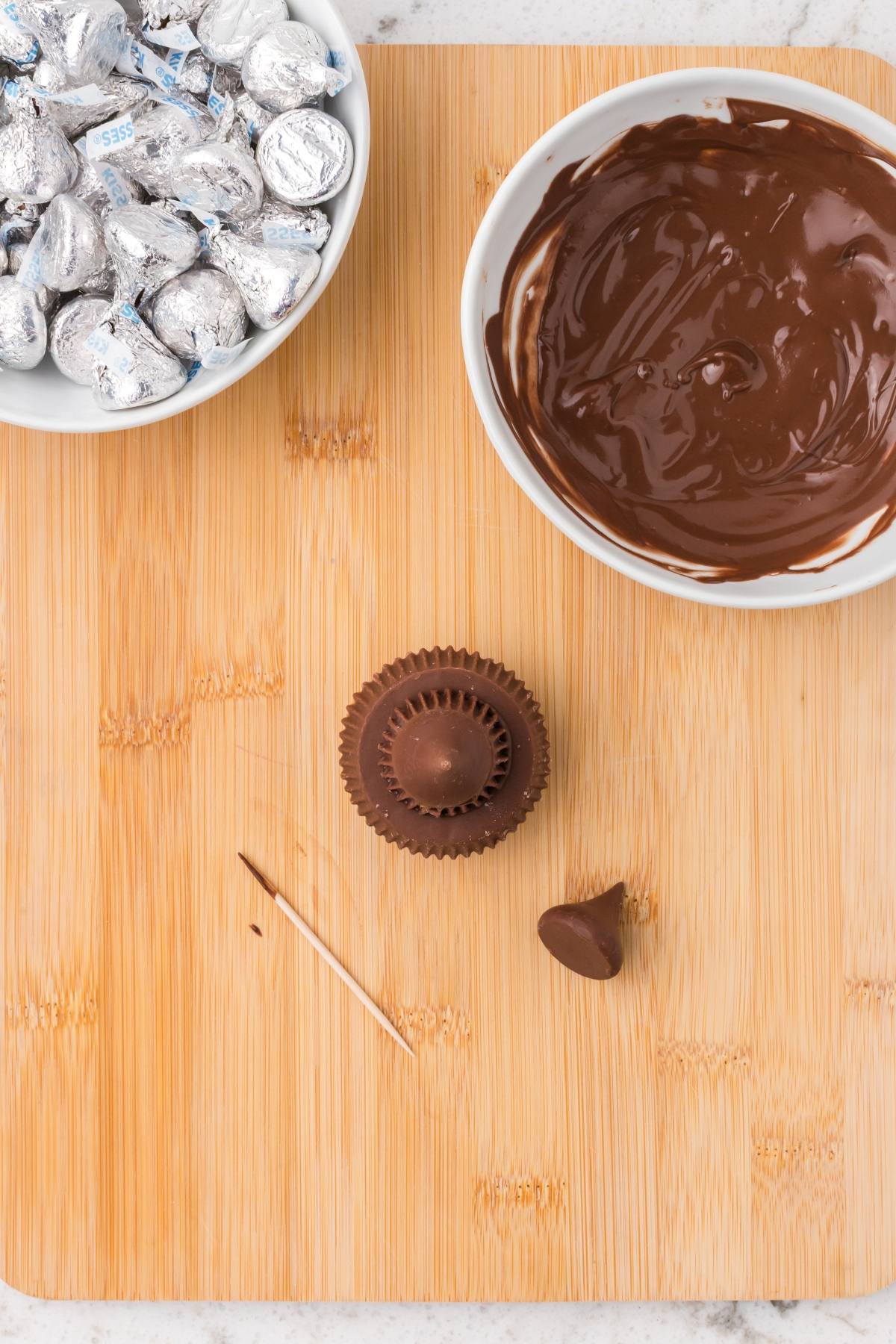 A wood board with a toothpick, chocolate Kiss, peanut butter cup, melted chocolate, and wrapped chocolate Kisses.