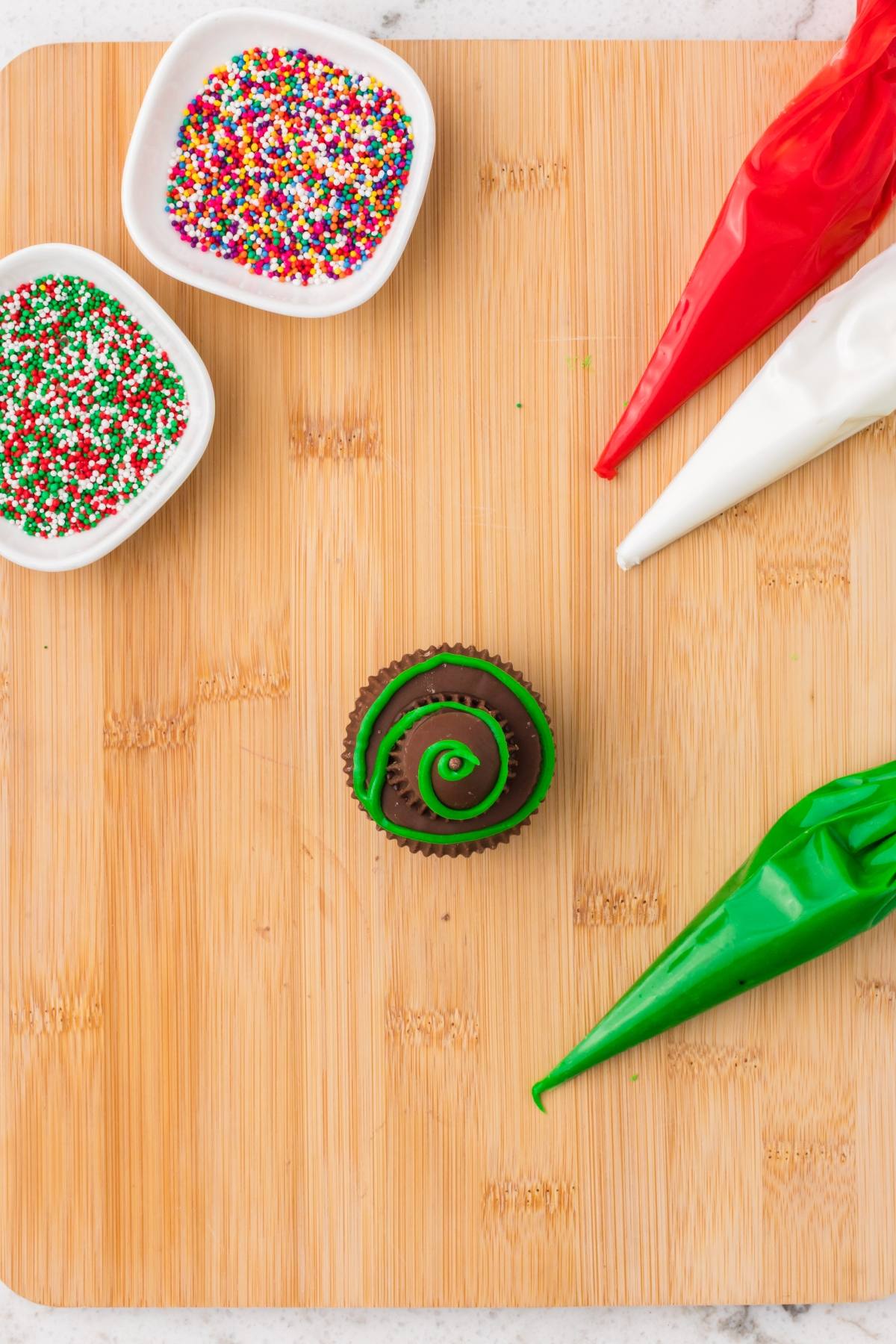 A cupcake with green icing, sprinkles, and red, white, and green frosting bags on a wooden board.