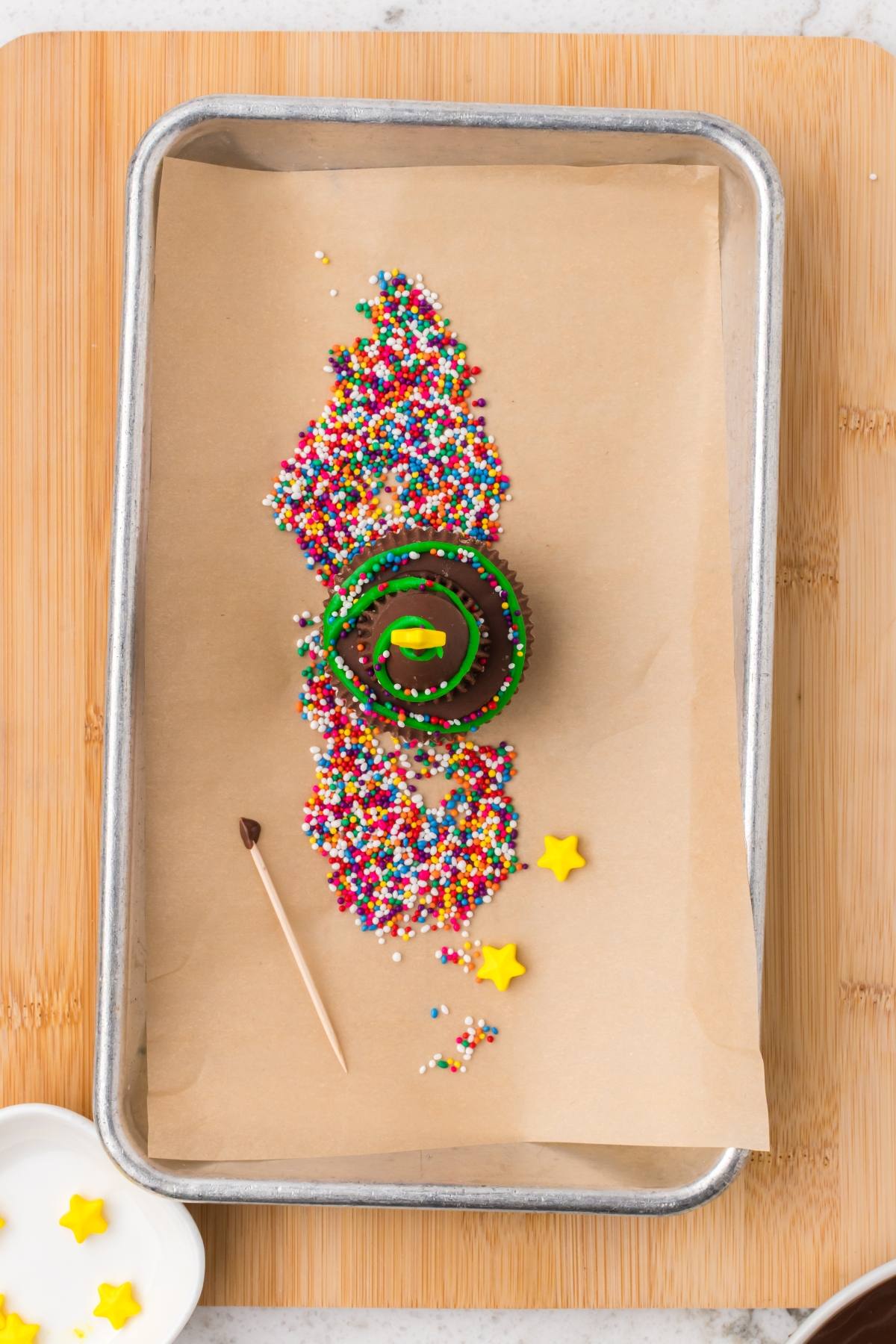 A chocolate cupcake with green icing and sprinkles on parchment paper, surrounded by colorful sprinkles and star candies.