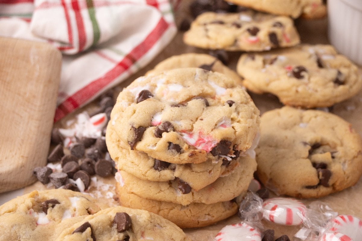 Peppermint Chocolate Chip Cookies with crushed peppermint pieces are stacked on a wooden surface, surrounded by festive candies and a striped towel nearby.