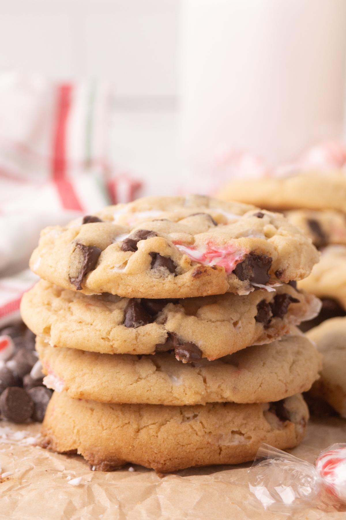 Four chocolate chip cookies stacked on parchment paper, with chocolate chips and peppermint pieces visible.