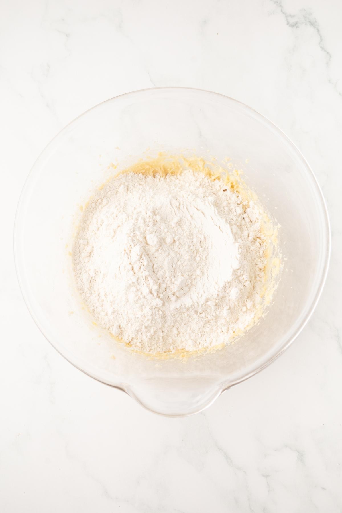 A glass bowl with flour on top of a yellowish mixture, placed on a white marble surface.