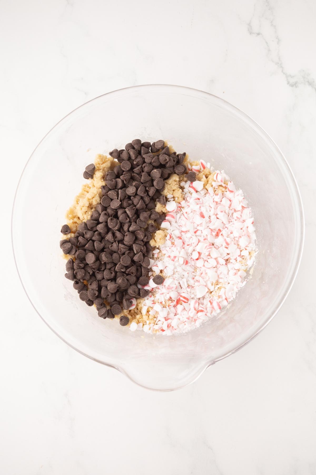 A glass bowl with chocolate chips, crushed peppermint, and cookie dough on a white marble surface.
