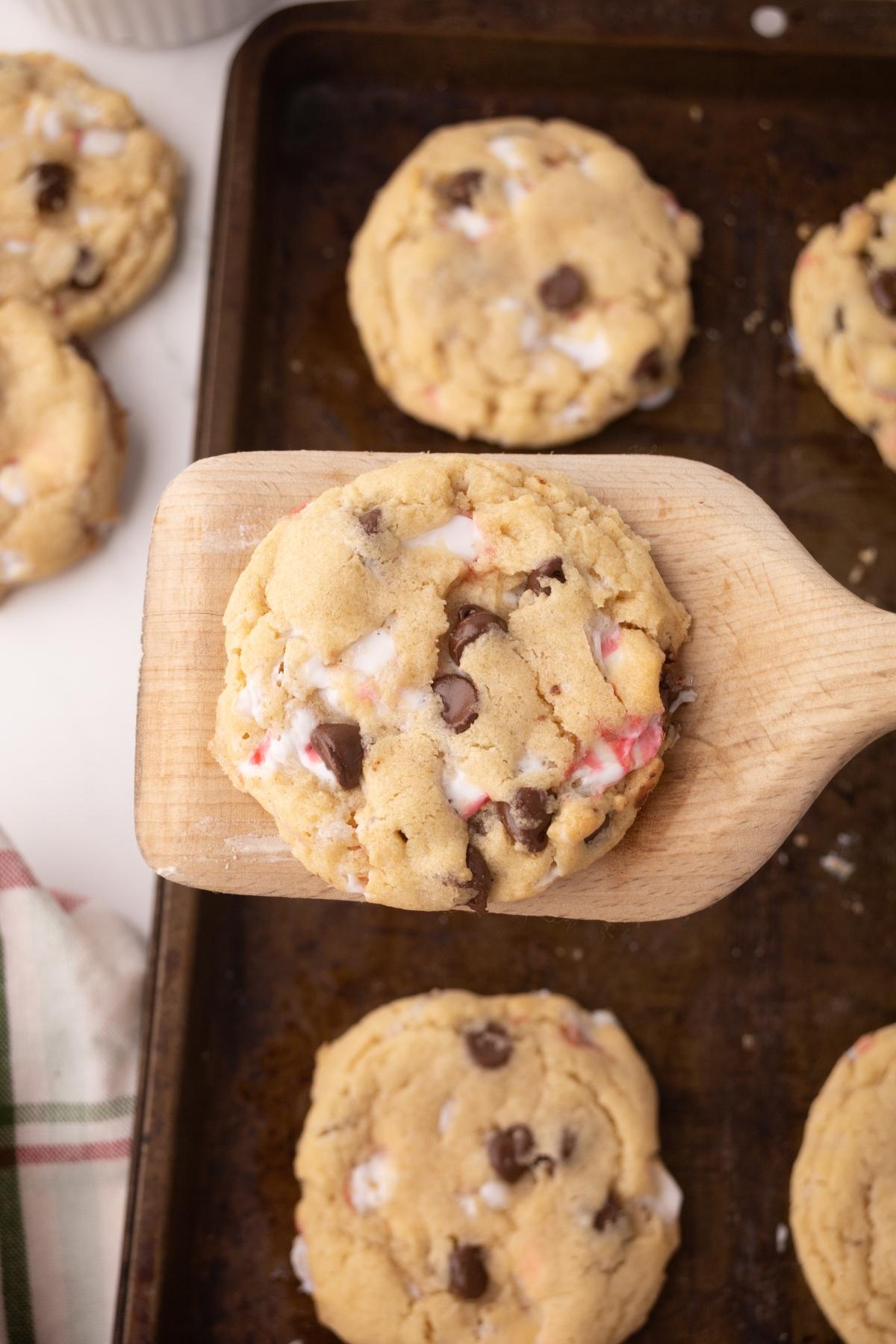 A peppermint chocolate chip cookie with marshmallows sits on a wooden spatula above a baking tray of cookies.